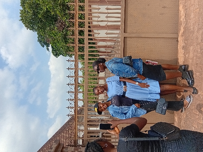 The image depicts four young adult females situated outdoors in Ejule, Nigeria, during daylight. On the left, one female, viewed from behind, is holding a black smartphone horizontally, directing it towards the other three subjects. She is wearing a black ribbed short-sleeved top and a matching skirt, with a black woven shoulder bag slung over her left shoulder. The three subjects are standing in a row, facing the camera, posing. The leftmost subject wears a black baseball cap, a light blue denim jacket over a black top, black trousers, and striped slip-on sandals, carrying a black quilted handbag. The central subject wears a light blue short-sleeved dress with a ruffled hem, cinched at the waist with a black belt featuring a gold-toned buckle, and black flat shoes, with a watch on her left wrist. The rightmost subject wears dark sunglasses, a light blue denim jacket over a black skirt, and black chunky-soled boots, carrying a small black shoulder bag. Two of the three posing subjects are smiling. Behind the individuals is a brown ornamental metal fence with scrollwork details and finials. Beyond the fence, to the left, a building with a reddish-brown tiled roof is visible, while to the right, a large green tree stands under a partly cloudy sky. The ground surface is paved with a reddish-brown hue.