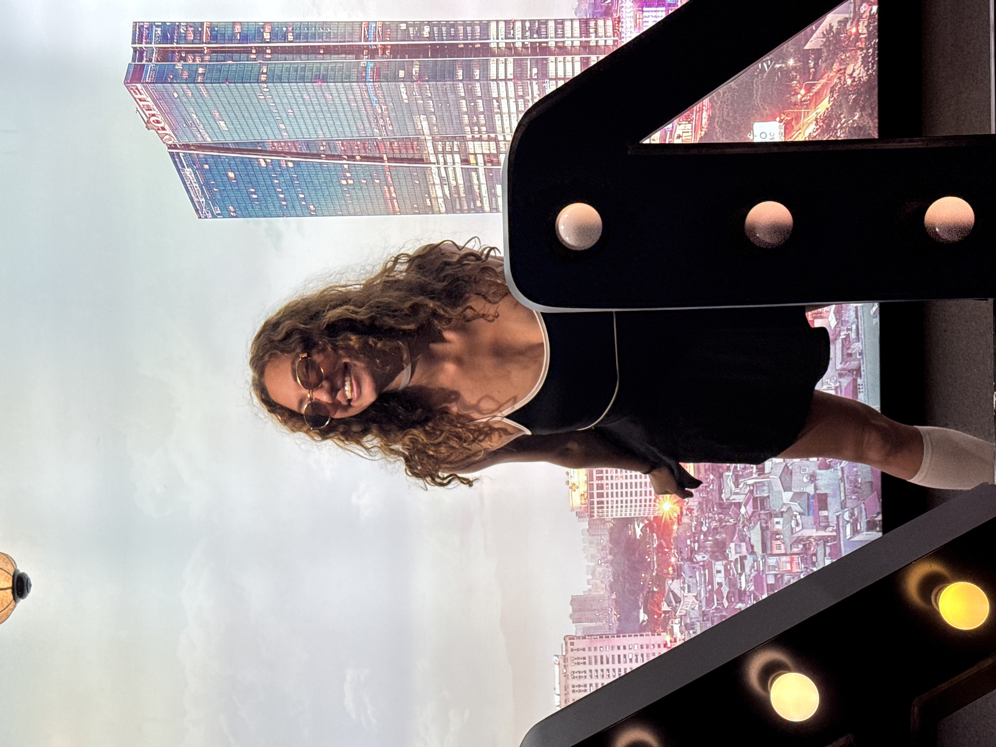 A woman with long, curly brown hair and sunglasses smiles widely, looking to her left. She is wearing a black dress with thin white straps and white thigh-high socks. She stands in front of a backdrop that displays a panoramic view of Hà Nội, Vietnam. The backdrop features several prominent buildings, including the Lotte Center Hanoi, a tall, modern skyscraper with a red "LOTTE" sign at its peak. The cityscape also includes numerous other buildings of varying heights and architectural styles, illuminated by city lights. In the foreground, on the left and right sides of the image, are large, black, decorative letter shapes with exposed light bulbs, obscuring parts of the backdrop. The sky in the backdrop is cloudy.