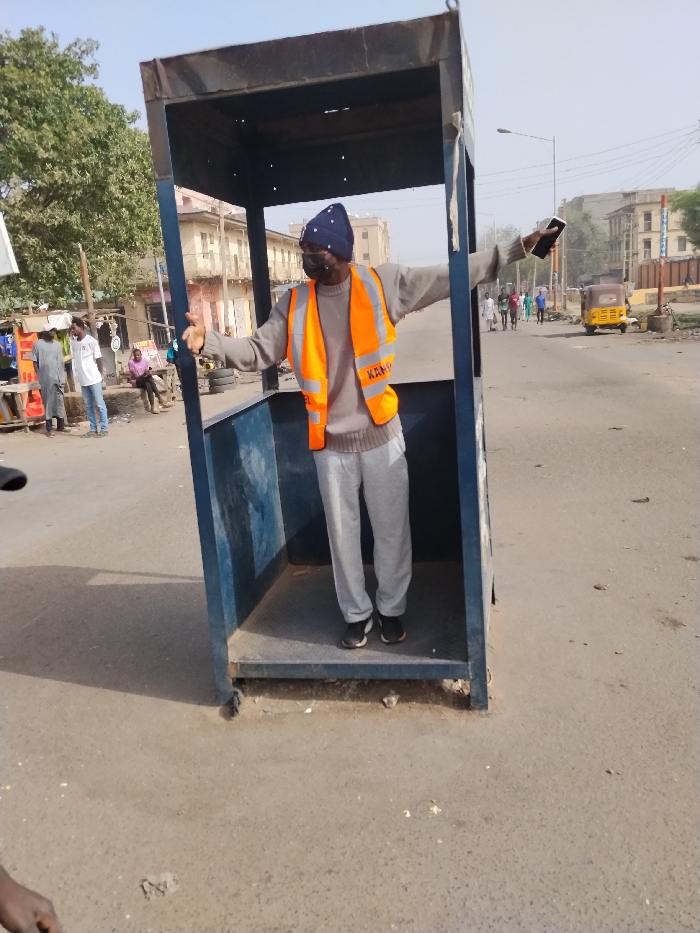 An outdoor forensic image captures a male individual standing centrally within a standalone, open-sided blue metal booth frame on an asphalt road in Sabon Gari East, Nigeria. The individual, of dark complexion, wears a dark blue knit beanie with white polka dots, a black face mask, a light grey long-sleeved sweater, an orange high-visibility vest with "KANO" printed in white on the left chest, light grey sweatpants, and black sneakers. He has his arms extended horizontally outwards, with a black smartphone held in his right hand.
To the left of the booth, several individuals are visible near a market-like setup with green foliage from trees behind it. Two men stand conversing, one in a grey garment, the other in a white t-shirt and blue jeans. A third man sits on a low structure in a pink shirt. Stacks of tires are visible on the left side of the road.
To the right of the booth, an asphalt road extends into the distance where multiple pedestrians walk. A yellow auto-rickshaw is present on the right shoulder of the road further down. Multi-story buildings with light-colored facades and a street light pole are visible in the background under a pale blue, hazy sky. The immediate foreground shows a partial view of a person's arm on the lower left. The road surface is a worn asphalt with scattered debris.