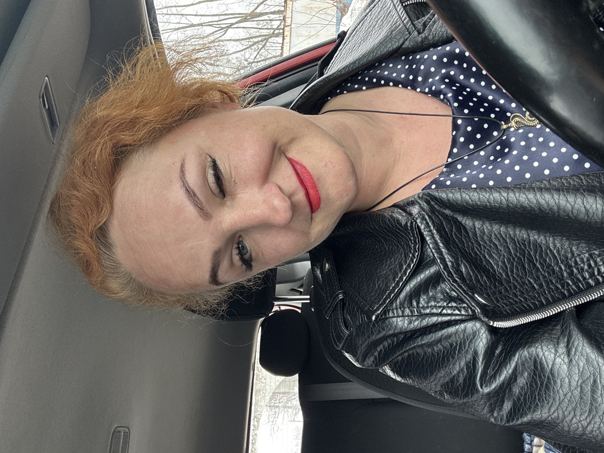A woman is in the driver's seat of a car. She is wearing a black leather jacket and a navy blue polka dot dress. She is also wearing red lipstick. She is smiling and winking. The photo was taken in Chaikovsky, Russia, which can be determined from the landscape visible through the car window.