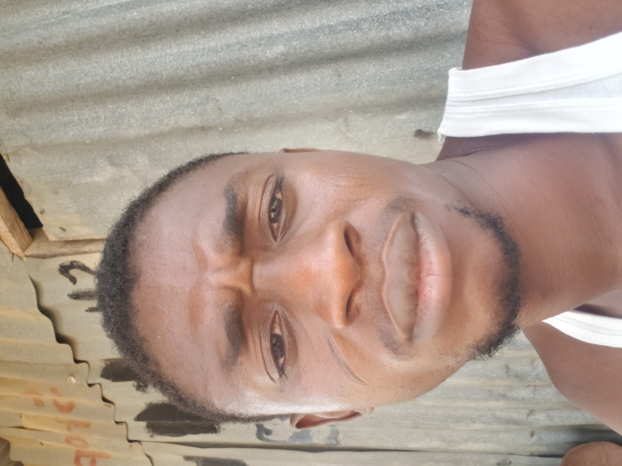 A close-up, rotated portrait features a young man of African descent looking directly into the camera with an intense and focused expression. His eyebrows are furrowed, his eyes are wide open, and his mouth is slightly ajar. He has short dark hair and a neatly trimmed goatee. He wears a white undershirt, visible on his right shoulder and chest (appearing in the upper right portion of the rotated image). The background consists of corrugated metal sheeting, likely a wall or roof, with faint, indistinct reddish markings visible on the lower left section. The lighting is bright and even, suggesting a daytime photograph. The setting appears to be an outdoor or semi-outdoor location, consistent with the provided context of Adankolo, Nigeria.