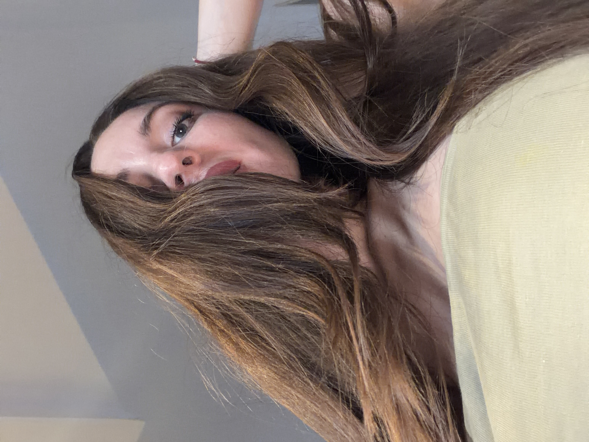 The image shows a close-up, low-angle shot of a person's face and hair, as if looking up from below. The subject has long, wavy, dark brown hair with lighter brown highlights, which falls across their face and obscures part of their features. Their eyes are open and looking towards the camera, with makeup visible on their eyelids and lashes. Their lips are slightly parted, revealing a hint of their teeth. The person is wearing a light green garment that covers their neck and shoulder, suggesting it might be a t-shirt or blouse. The background is a plain, light gray wall, with a shadow cast across it from an unseen light source. The overall lighting is soft, with no indication of the time of day or specific weather conditions. The scene appears to be indoors, possibly in a bedroom or a casually decorated room. There is no visible text in the image. The cropping of the image focuses intensely on the person's face and hair, creating an intimate and slightly dramatic effect.