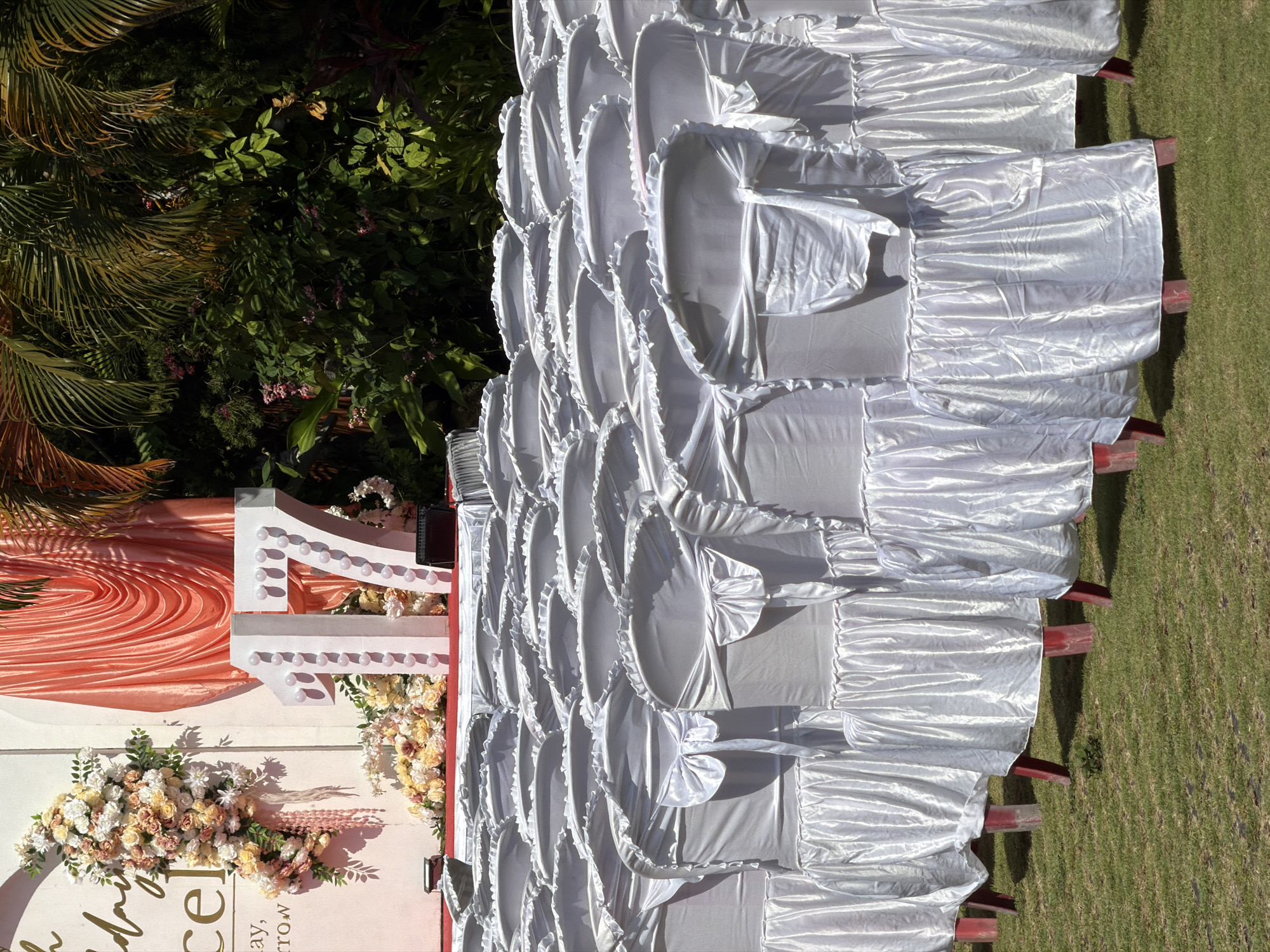 This is a medium shot, taken outdoors in daylight, of a row of white-covered chairs set up on a green lawn. The chairs are arranged in multiple rows, with the closest row in sharp focus and the rows behind receding into a soft blur. Each chair is covered with a white fabric that drapes over the back and seat, tied with a large white bow on the back. The fabric has a shiny, satin-like finish and is ruffled at the edges. The legs of the chairs are visible and appear to be red.
In the background, there is a decorated wall or backdrop. To the left of the backdrop, there is a large floral arrangement in shades of cream, peach, and pink, with green foliage. To the right of the floral arrangement, there is gold lettering on the backdrop, partially visible. A large number "17" made of white material and lit with bulbs is prominent in the center of the backdrop, flanked by more floral arrangements. Behind the number "17", there are draped coral-colored fabrics. To the far right of the image, lush green foliage, including palm fronds, is visible.
The scene appears to be set up for an event, possibly a party or celebration, given the decorations and the formal arrangement of chairs. The presence of palm trees and dense foliage suggests a tropical or subtropical location.
While the image does not explicitly show a specific location in North Kuta, Indonesia, the lush greenery and the style of outdoor event setup are consistent with locations in that region.