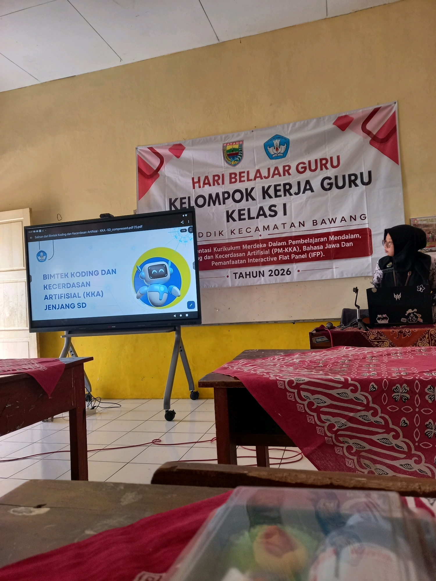 The image captures an indoor scene, likely a classroom or meeting room, in Sangubanyu, Indonesia. A large banner is prominently displayed, featuring the text "HARI BELAJAR GURU KELOMPOK KERJA GURU KELAS I" followed by "DIKDIK KECAMATAN BAWANG". Below this, further text details the agenda: "entasi Kurikulum Merdeka Dalam Pembelajaran Mendalam, ng dan Kecerdasan Artificial (PM-KKA), Bahasa Jawa Dan Pemanfaatan Interactive Flat Panel (IFP). TAHUN 2026".
In the foreground, a large monitor displays a presentation with the title "BIMTEK KODING DAN KECERDASAN ARTIFISIAL (KKA) JENJANG SD", accompanied by a cartoon robot graphic. Two wooden tables are visible, draped with red cloths featuring a patterned design.
To the right, a woman wearing a headscarf and glasses is seated at a table, looking towards the monitor and holding a laptop. A black laptop is open in front of her, adorned with stickers. The room has a yellow-painted wall and a tiled floor. Electrical cords are visible on the floor. The overall ambiance suggests an educational event or workshop.