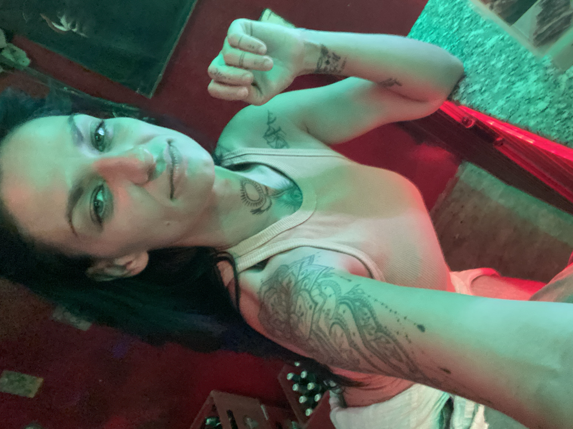 An adult female is depicted in a selfie taken indoors, likely a bar, in Mũi Né, Vietnam. She has long, dark hair, a slight smile, and gazes directly into the camera. She wears a light-colored ribbed tank top, revealing multiple tattoos: a large, intricate design covering her left upper arm, a circular motif on her neck, a mountainous design on her right upper arm, and an abstract mark on her right wrist. Her right hand is observed in a clenched fist position. The subject leans on a counter featuring a speckled green surface and an integrated red light strip along its edge. The background consists of a red wall, with two wooden crates containing bottles visible on the left. The scene is illuminated by strong artificial green and red lights, resulting in distinct color casts across the subject and surroundings.