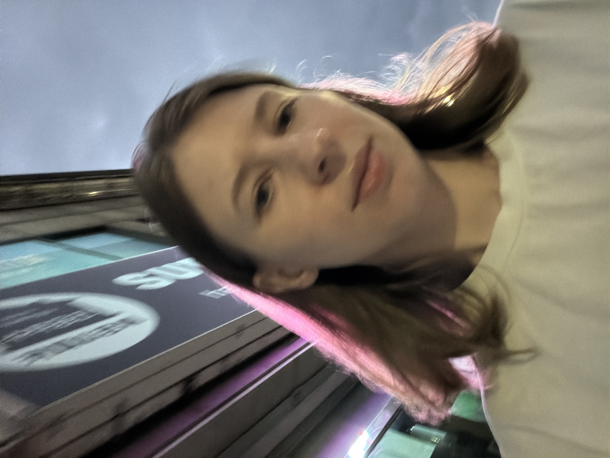 The image shows a young woman from the chest up, looking towards the camera. She has light brown hair, some of which is illuminated with a pink hue from a nearby light source. She is wearing a plain white t-shirt. The background is blurry, but appears to be an urban environment with a sign that has the letter "S" and a circular graphic visible. The overall lighting suggests it is either dusk or dawn, or an indoor setting with artificial lighting. The image was captured outdoors in the city of Dongducheon-si, South Korea.