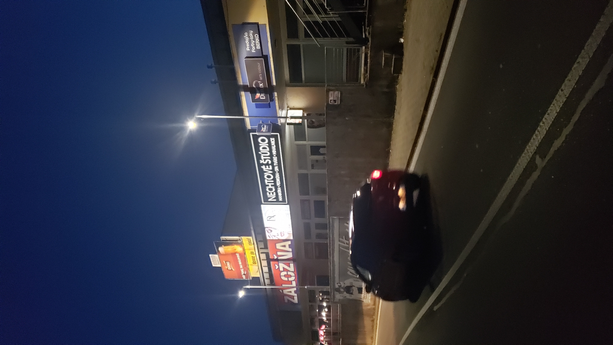 The image shows a street scene at dusk or night in Prešov, Slovakia, with a building featuring several illuminated signs and a dark-colored car driving on the road in the foreground. The building's facade has a concrete finish. Prominent signs include "ZÁLOŽNA" (which translates to "Pawnshop"), "NECHTOVÉ ŠTÚDIO" (which translates to "Nail Studio"), and a sign for "Predajňa Farby-laky BERKO" (which translates to "Paint and Varnish Store BERKO"). Another sign depicts a glass of beer and text related to breakfast. A street lamp is visible in the upper right portion of the image. The car in the foreground is blurred due to motion.