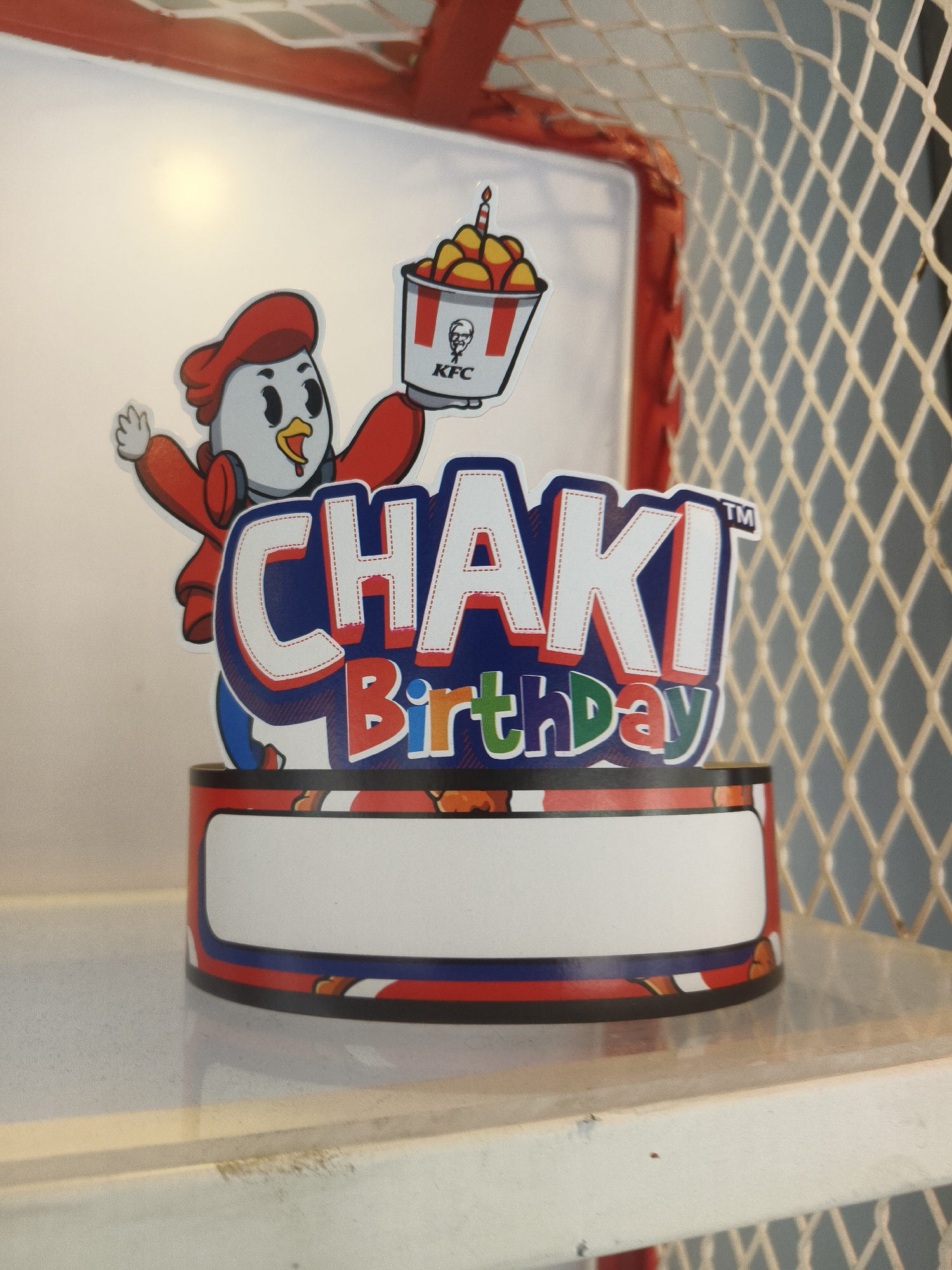 An outdoor promotional display features a cartoon chicken character wearing a red hat and jacket, holding a bucket of fried chicken with a lit candle. The character stands on a cylindrical base decorated with red, white, and blue stripes. The words "CHAKI Birthday" are prominently displayed on the base. The scene appears to be set up for an event or promotion in a public area, possibly at a KFC restaurant. The background shows a white surface, a red structure, and a cream-colored netting. The lighting is bright, suggesting daylight. The location is identified as Denpasar, Indonesia.