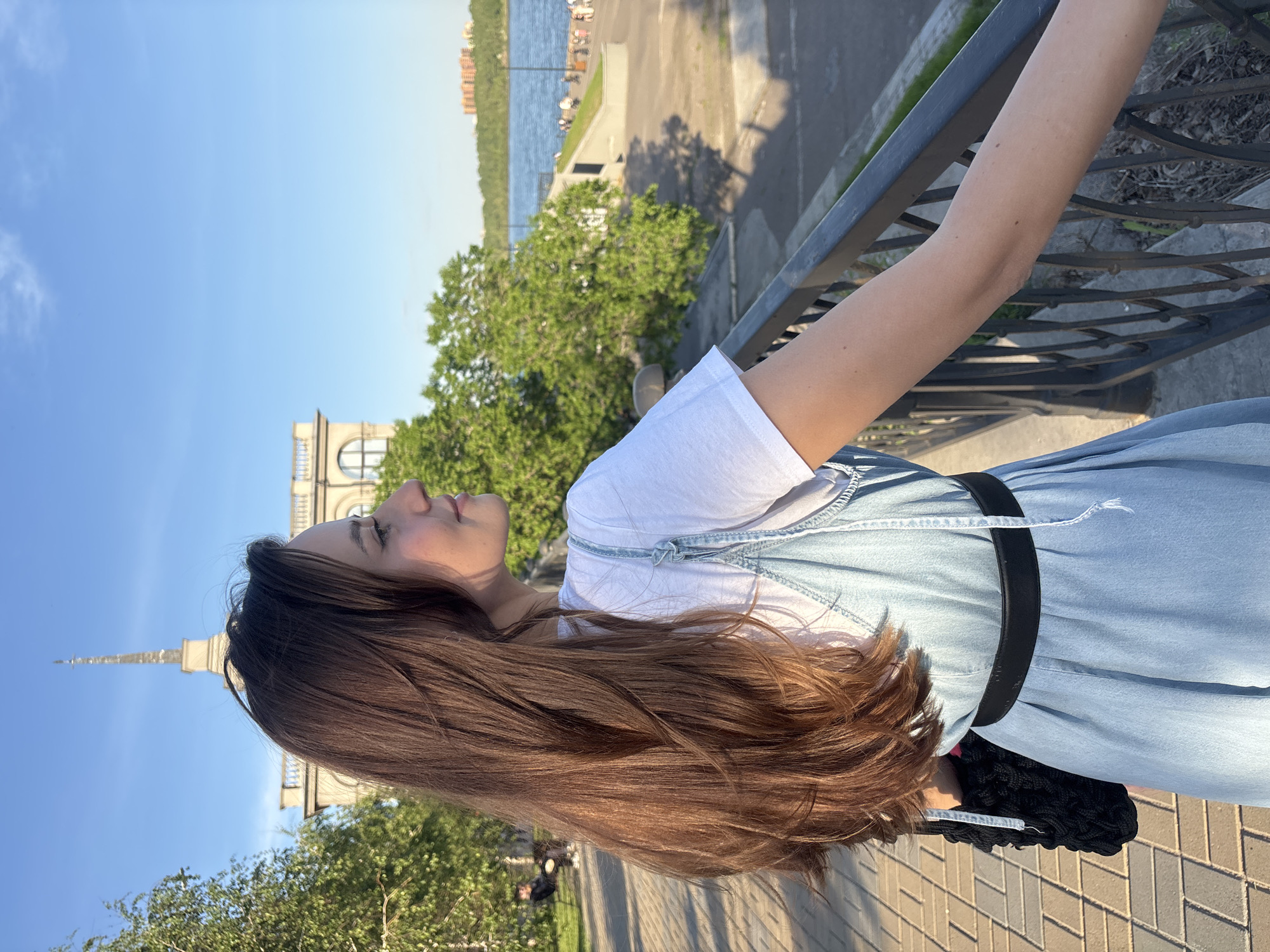 The image, originally oriented sideways, depicts a young woman standing outdoors on a sunny day in Krasnoyarsk, Russia. She is looking upwards and slightly to her left (the viewer's right) with a serene expression, a faint smile gracing her lips. Her long, wavy brown hair cascades down her back and shoulder, warmly illuminated by the sun. She wears a white t-shirt underneath a light blue, denim-like pinafore dress, cinched with a black belt. Her right arm rests casually on a black metal railing that runs along the foreground.
The background features a clear blue sky with a few wispy clouds. A notable light-colored building with classical architectural details, including arched windows and a distinctive, ornate spire, is visible beyond her, partially obscured by green trees. Further in the distance, a wide body of water, characteristic of the Yenisei River, stretches across the horizon, with some structures visible on the opposite bank. The setting appears to be a riverside promenade or park with a paved walkway. The warm, golden light suggests it is late afternoon or early evening.