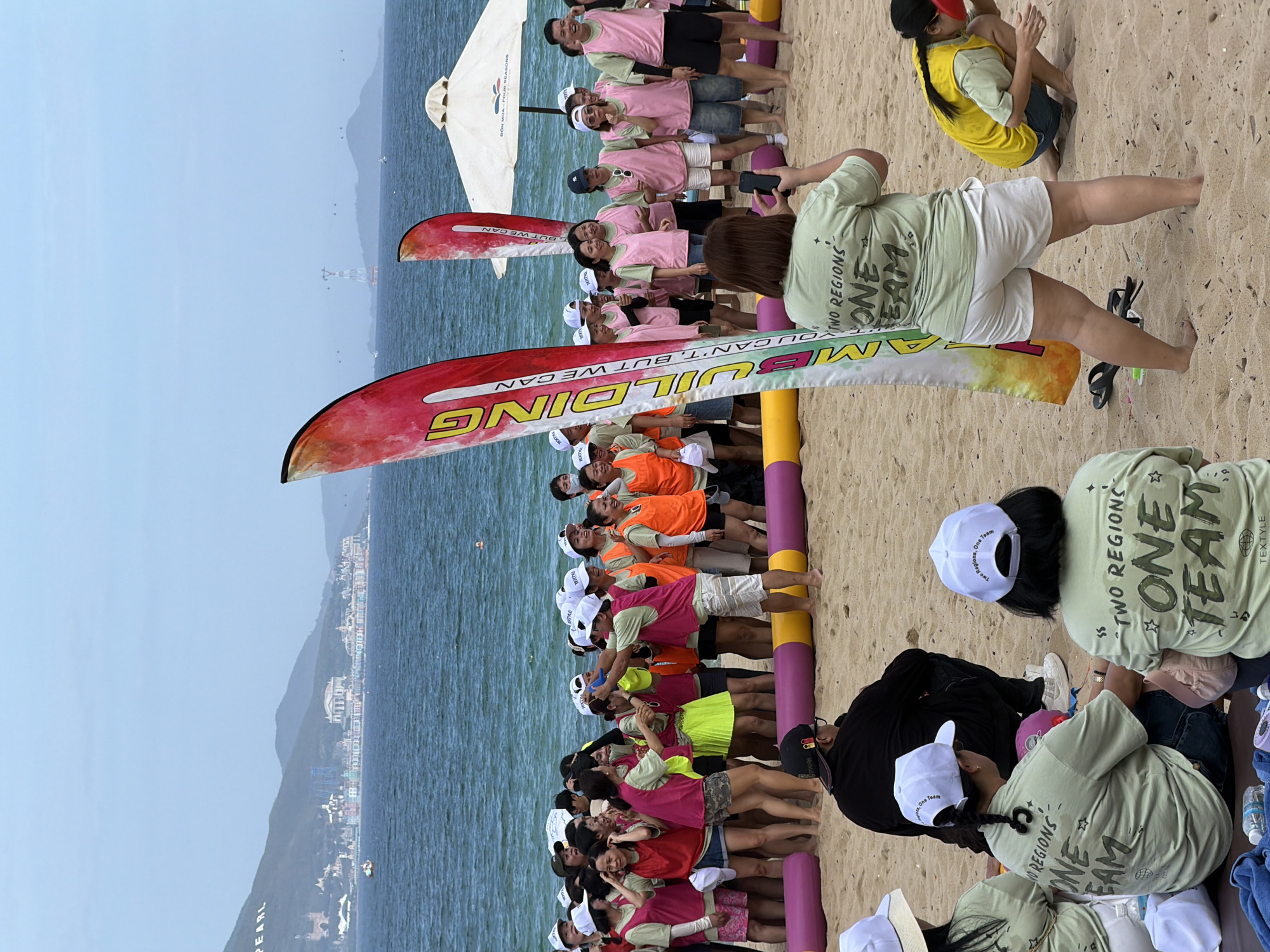 A group of people are gathered on a sandy beach in Nha Trang, Vietnam, with the ocean and a city skyline in the background. The sky is overcast. In the foreground, several individuals are wearing light green t-shirts with the text "TWO REGIONS ONE TEAM" printed on the back. Some are wearing white baseball caps. A large banner with the word "BUILDING" printed vertically and "BUT WE CAN" horizontally is visible. Several people are interacting, with one woman in a pink vest appearing to point a blue object at another person. Two large inflatable tubes, one purple and one yellow, are placed horizontally on the sand. In the middle ground, a large group of people are wearing either pink or orange vests over their t-shirts, and many are wearing white hats. A white umbrella is set up on the beach. In the background, a coastal city with buildings and mountains is visible. The ocean stretches out to the horizon.