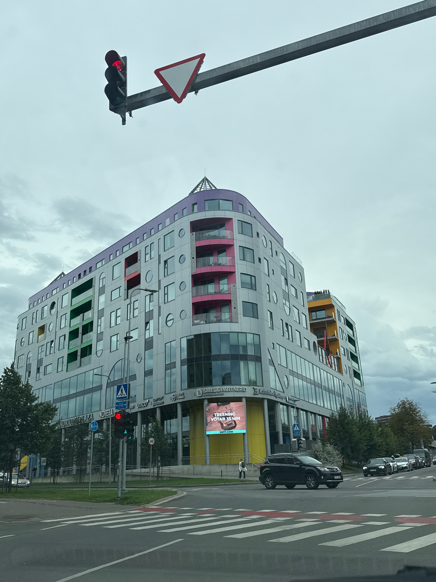 The image captures a street-level view of a modern, multi-story building with a colorful, undulating facade, featuring balconies with pink and green accents, and circular window elements. The building's signage indicates it is located in Tallinn, Estonia, specifically at "ADRICHI PLAZA" and "NOVELLA". A traffic light displaying a red signal is prominently visible in the foreground, suspended over the intersection. Adjacent to the traffic light is a triangular yield sign. Several cars are stopped at the intersection, waiting for the light to change. A pedestrian is visible on the sidewalk in front of the building. The sky is overcast, suggesting a cloudy day. The foreground shows a crosswalk and road markings. The overall scene depicts an urban environment with functional traffic infrastructure and contemporary architecture.