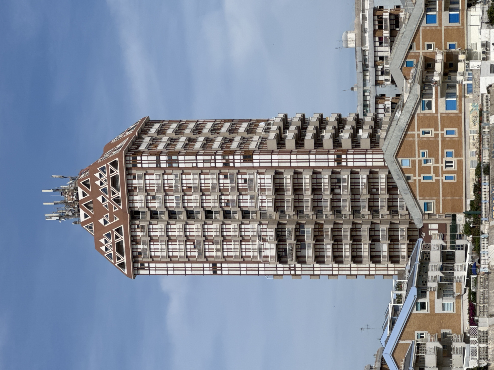 The image showcases the Torre Palazzi, a prominent high-rise building located in Nettuno, Italy. The tower is distinguished by its unique architectural design, featuring a dark brown angular peak adorned with triangular shapes and an array of telecommunications antennas at its apex. The building's facade is characterized by repeating patterns of rectangular windows and balconies, creating a grid-like structure. Adjacent to the tower is a complex of lower-rise buildings of similar style with a terraced design. The sky above is clear and blue, offering a bright background. The photograph seems to have been rotated 90 degrees counter-clockwise.