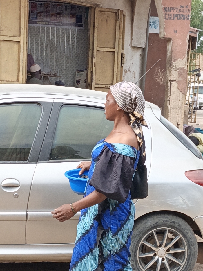 The scene is set in Gwagwarwa, Nigeria. An adult female, positioned centrally and in profile facing left, walks along an unpaved surface next to a parked silver Peugeot 206 sedan. She wears a patterned headscarf, a blue and teal off-the-shoulder dress with black puffed sleeves, and carries a small, dark shoulder bag. Her right hand holds a bright blue plastic bowl containing several dark, irregularly shaped objects. Her mouth is slightly open. A light-colored bracelet adorns her right wrist.
To her left, partially obscured by the car, is an open storefront with aged, light-brown wooden double doors. Inside, a person wearing a light-colored cap is seated, facing away from the camera, with shelves displaying packaged goods visible on the back wall. Above the storefront, faded signage is visible.
To the right of the woman and car, a deteriorated reddish-brown building facade features peeling paint and partial signage reading "HAPPY SALLA" and "CALIFORNIA." A thin metal antenna protrudes from this wall, and a metal structure resembling scaffolding or a ladder leans against it further right. In the far background, additional indistinct figures and vehicles are visible, including one individual lying or crouching near the ground to the right of the parked car. The ground appears dusty and unpaved. The scene is illuminated by natural daylight.