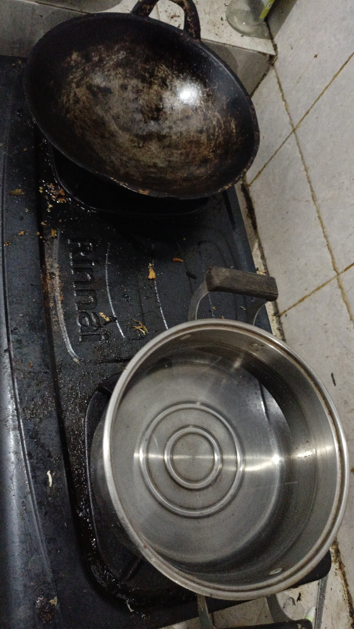 A close-up, high-angle shot shows a greasy, black gas stove with a dark, well-used wok placed on top. In front of the wok, a stainless steel pot with a concentric circular pattern on its interior sits on the stove. The stove surface and surrounding tiled wall appear dirty, with visible food debris and grease. The image is taken indoors, likely in a kitchen, and there are no people or animals present. The location is identified as Trihanggo, Indonesia.