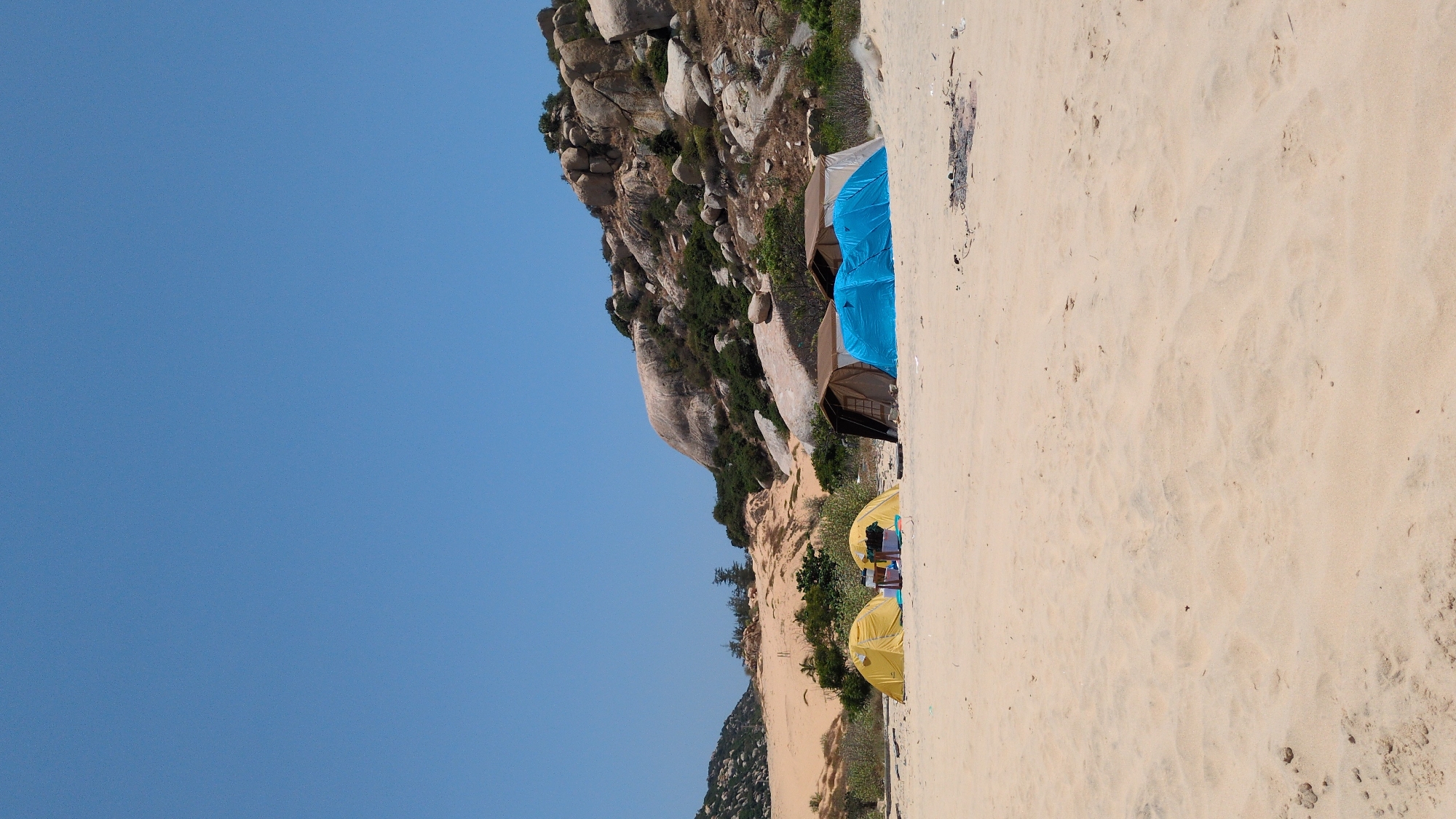 This image depicts a beachside camping area. In Phuoc Dinh, Vietnam, a series of tents are set up against a backdrop of rocky hills and a sand dune. The tents vary in color; some are blue and brown, and others are yellow. The environment consists of sand, rocks, sparse vegetation, and a clear blue sky. There are no identifiable people visible, but the tents suggest occupancy. An area of disturbed sand, potentially indicating a previous fire, is present near the tents.