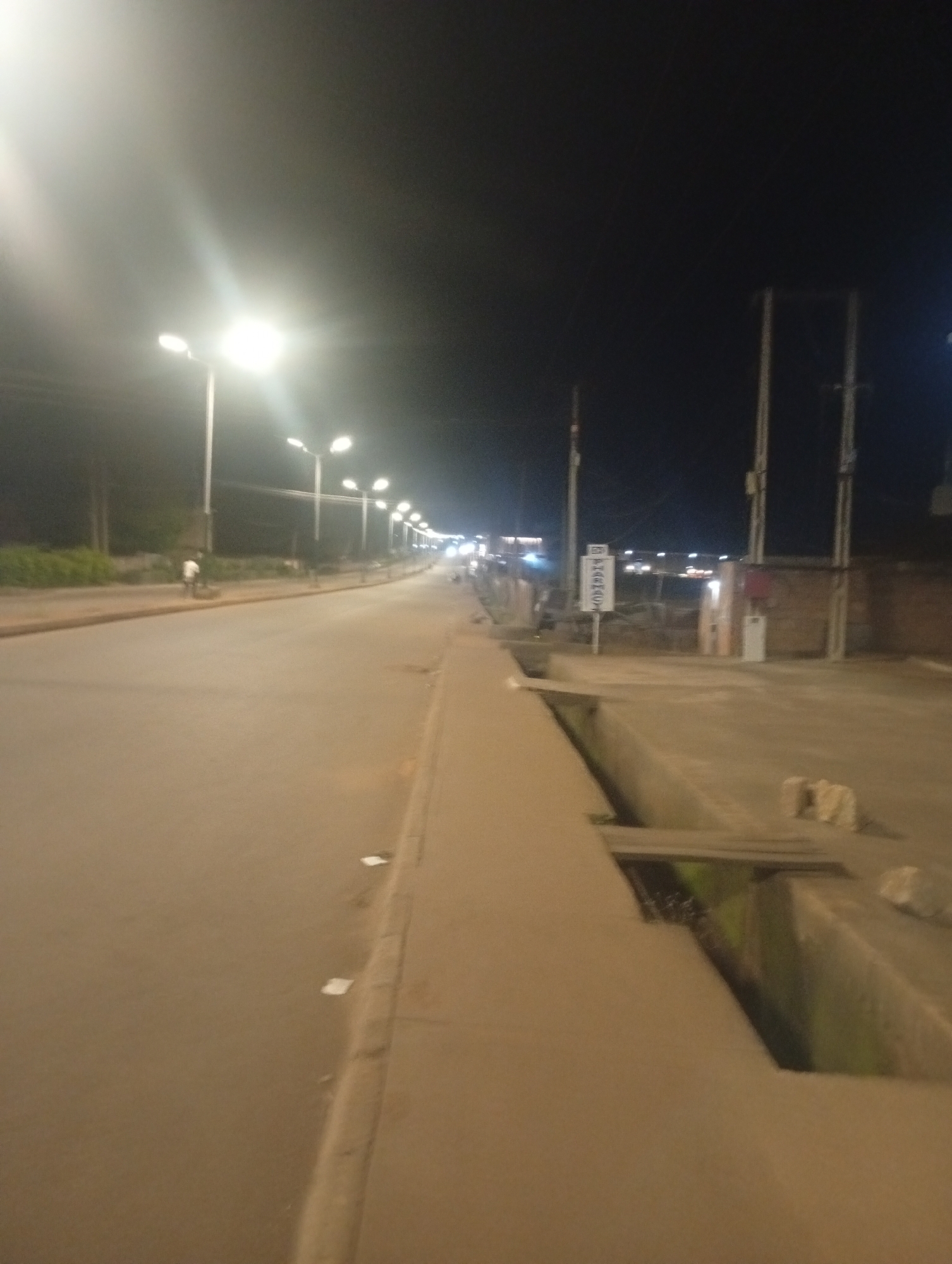 The image depicts a street scene at night in Bukuru, Nigeria. A street light illuminates the asphalt road, which curves to the right. Several other street lights are visible along the road, stretching into the distance. On the left side of the road, there is a sidewalk with vegetation and a lone figure walking. On the right side of the road, there is a concrete drain, a brick wall, and a sign that reads "BULU." The overall scene is dimly lit, with the street lights providing the primary source of illumination.