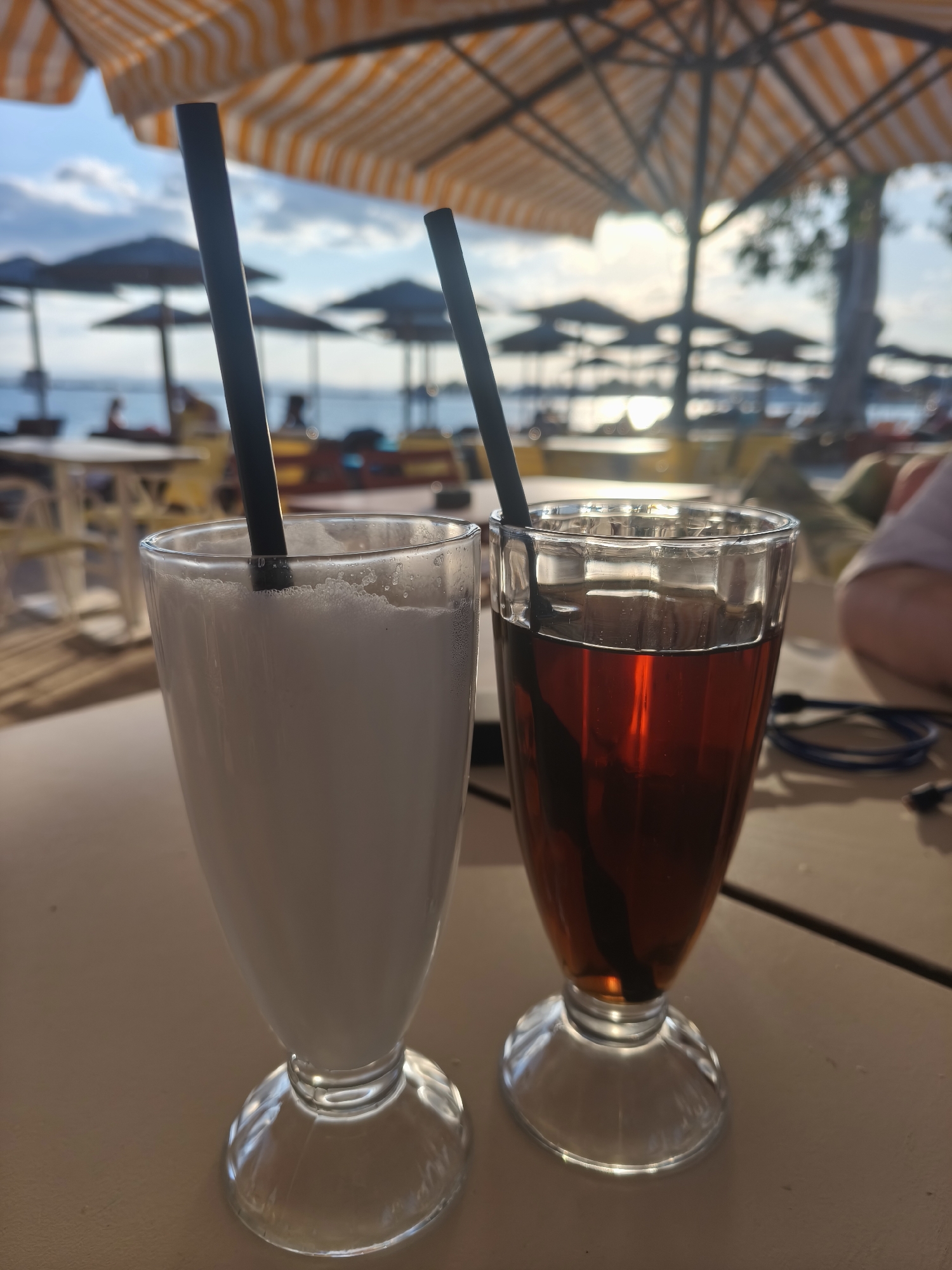 Two glasses of drinks, one white and frothy, and the other a dark amber liquid, stand on a table. Both glasses have black straws. In the background, beach umbrellas are set up on a beach. The scene appears to be outdoors, possibly at a beachside cafe in Eretria Municipal Unit, Greece. The lighting suggests it is daytime, with the sun casting a warm glow. There are glimpses of people in the background, some seated at tables and others near the water. The overall atmosphere is relaxed and indicative of a vacation or leisure setting.