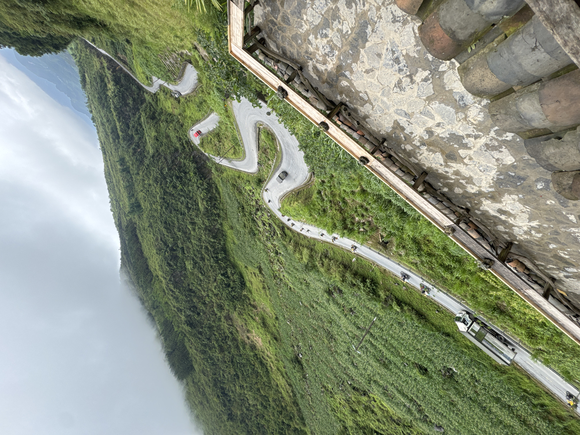 Captured from a high vantage point adjacent to a building with a tile and stone roof, the scene depicts a winding mountain road in Yên Minh Commune, Vietnam. The road traverses a steep, verdant slope, densely covered in vegetation, descending into a foggy valley. Several vehicles, including cars and a truck, are visible on the road, along with a group of cyclists. The road features sharp hairpin turns as it ascends the mountain. The overall perspective offers a panoramic view of the mountainous landscape and the road infrastructure cutting through it.
