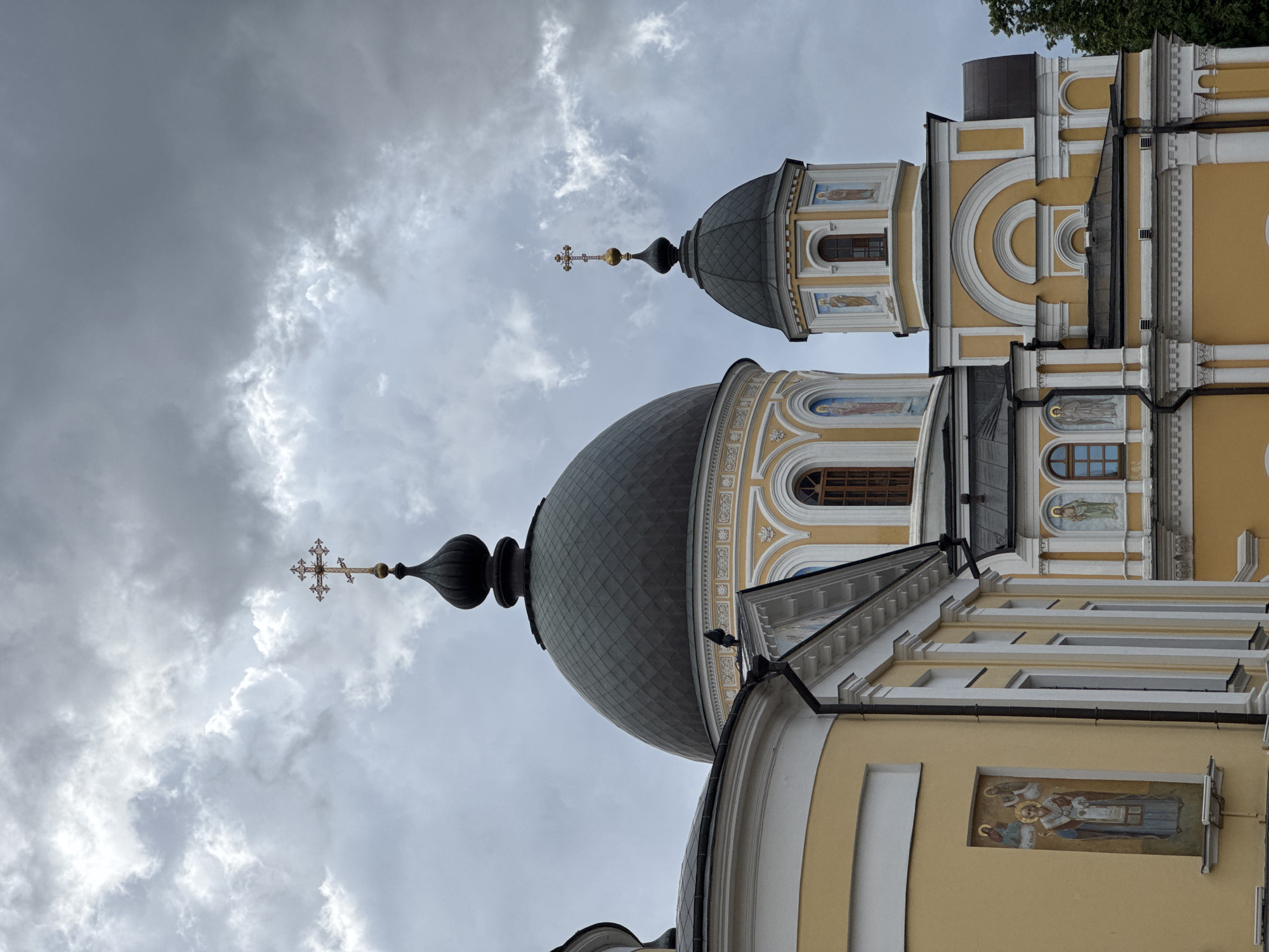 The image depicts a portion of the Danilov Monastery in Moscow, Russia. The focal point is on the monastery's architectural details: the dome-shaped roofs topped with Orthodox crosses, and the yellow and white facade with religious artwork. The sky is overcast with heavy, grey clouds.