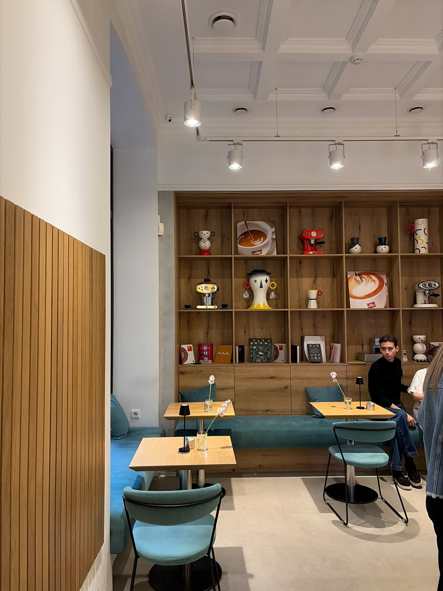 The image depicts the interior of a cafe, likely located in Minsk, Belarus. The scene is bright and modern, with light-colored walls and a coffered ceiling. To the left, a section of a wall is covered in vertical wooden slats, creating a warm textural element.
In the center of the frame, a tall wooden shelving unit is filled with various decorative items, including what appear to be coffee-related memorabilia, small appliances resembling espresso machines, vases, and framed artwork. The shelves are illuminated by the overall lighting of the cafe.
In the foreground and mid-ground, there are several light-colored wooden tables with minimalist black bases. Two tables are visible in detail, each set with a small black lamp and a clear glass containing water and a single flower. One table has a teal cushion on the bench behind it.
A teal upholstered bench runs along the wall beneath the shelving unit, providing seating. Two light blue chairs with black metal frames are positioned at the tables.
In the background, to the right, a young man wearing a black sweater and jeans is seated at one of the tables, looking towards the left. Another person, whose body is partially visible from the shoulder down and wearing a denim jacket, is standing to the far right.
The overall ambiance suggests a contemporary coffee shop or cafe, possibly a chain, given the uniformity of some decorative items. The flooring appears to be a light gray, polished concrete or similar material.