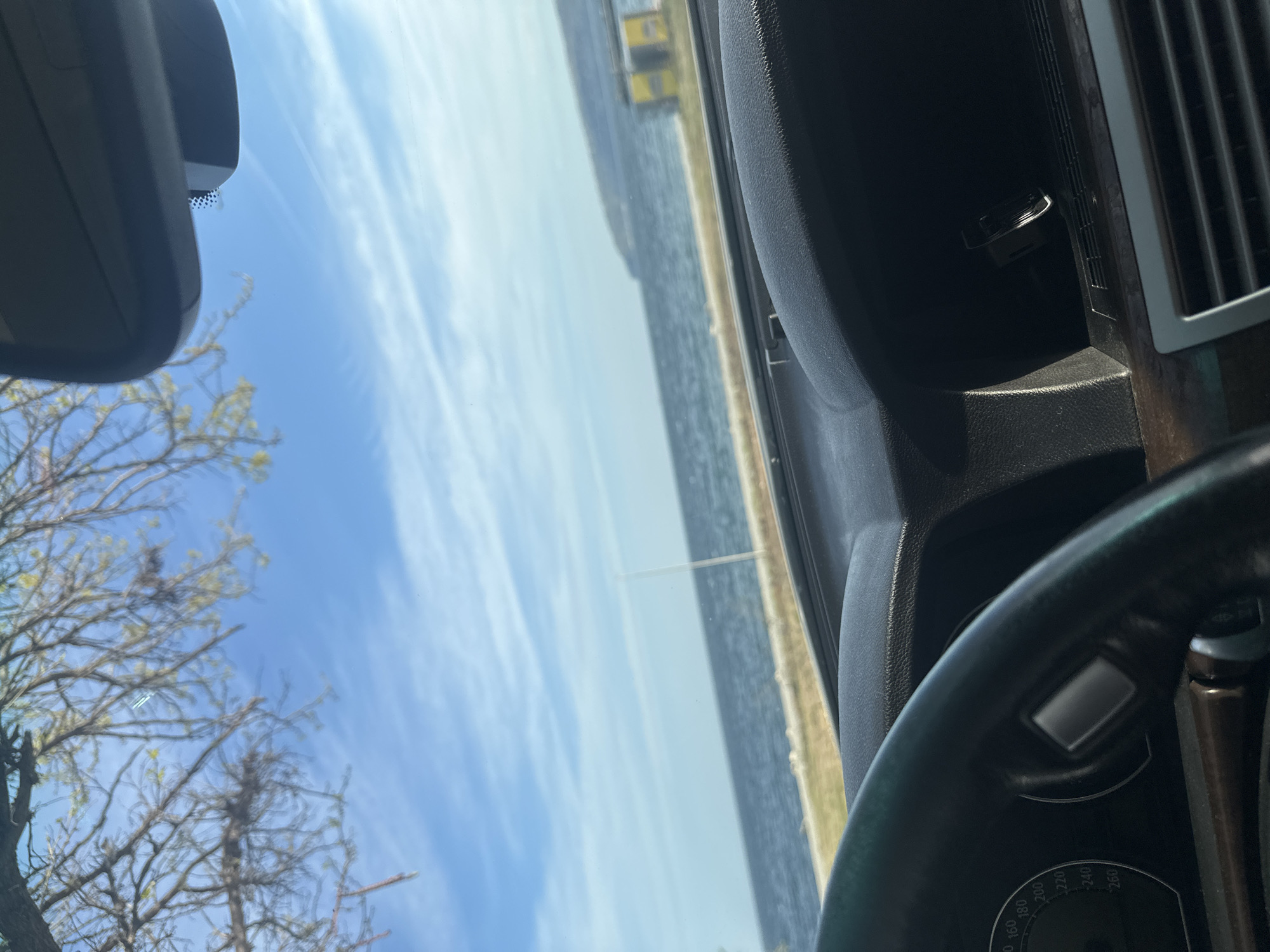 The image presents a daytime view from inside a vehicle, looking out through the front windshield. The scene captures a blend of car interior, nature, and a large body of water.
In the foreground, sections of the car's interior are visible. On the lower right, a black steering wheel partially covers a gauge cluster, where speedometer markings from 160 to 260 (likely km/h) are discernable. A dark dashboard with subtle wood-grain accents features a chrome-trimmed air vent, and a USB charger is plugged into a port. At the top center, the base of a rear-view mirror is affixed to the windshield, against a dotted or textured area of glass.
Through the windshield, a vast expanse of calm, blue water stretches across the middle ground, indicating a sea or large lake. A faint, distant shoreline or landmass can be seen, with some indistinct yellow structures. Above the water, the sky is a clear blue with scattered wispy white clouds, suggesting sunny weather. To the far left, a network of bare tree branches with emergent green leaves frames the view of the sky, hinting at springtime.
The image conveys a peaceful atmosphere, likely taken while parked or driving slowly alongside the water, consistent with a coastal setting like Varna, Bulgaria. No individuals are visible in the frame.