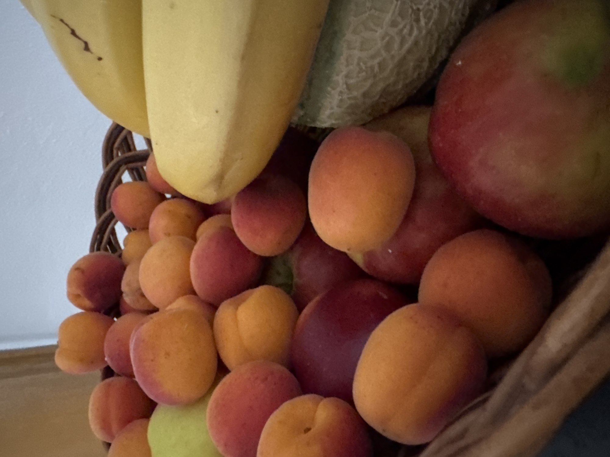 The image depicts a close-up view of a wicker basket filled with a variety of fruits. Prominently featured are apricots, their skins showing a gradient of orange and red hues. A few bananas are visible at the top, with their characteristic yellow color. Partially obscured by the fruits, the textured rind of a melon can be seen. The background is a soft, diffused light, suggesting an indoor setting. The overall impression is one of abundant, fresh produce, likely a home pantry or a market stall in Hohenbrunn, Germany.