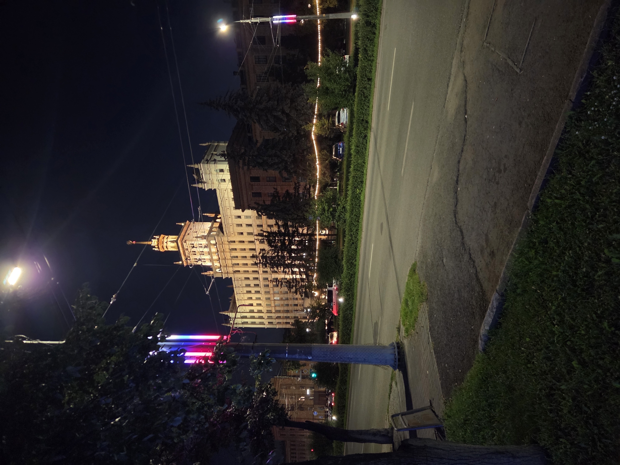 A wide, multi-lane road runs through the foreground, with cars visible in the distance. On the left, a tree and a lamppost with colorful lights are prominent. The lamppost has a blue, white, and red light display, possibly the Russian flag colors. In the background, a large, illuminated building with a tower stands out against the night sky, characteristic of Stalinist architecture. This building is identifiable as the Main Building of South Ural State University (SUSU) in Chelyabinsk, Russia. The area appears to be an urban environment with trees and decorative lights along the street.