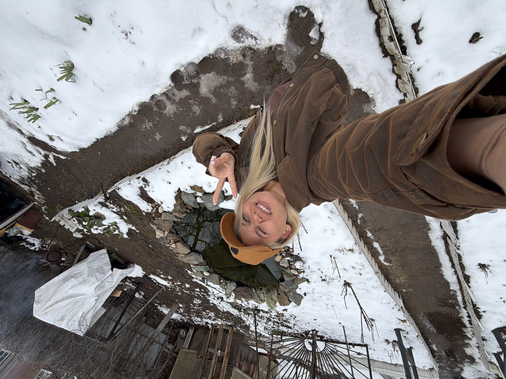 A blonde woman in a brown jacket and cap is taking a selfie in a snowy outdoor location in Sarapul, Russia. She is holding up a peace sign with her left hand. Behind her, there is a small pond surrounded by stones. The ground is covered with snow, and there are various objects visible, including a white tarp, a metal structure, and some plants. The picture is taken from a high angle.