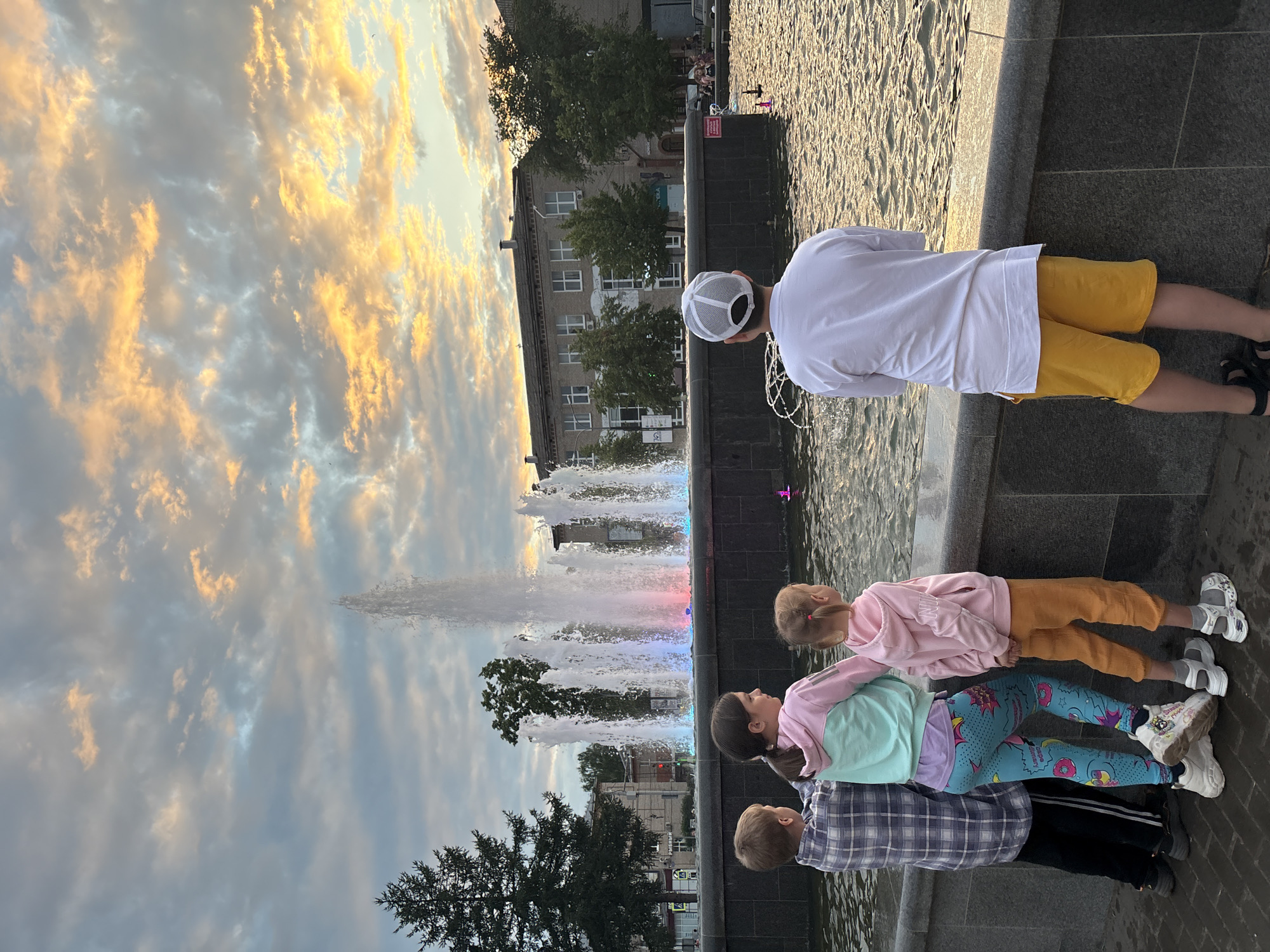 In the city of Chaikovsky, Russia, four children stand by a fountain, their backs to the camera, observing the water jets illuminated with colored lights. The sky above is filled with dramatic, colorful clouds, catching the last light of day. The fountain is central to the scene, with its powerful streams of water creating a dynamic visual element. Behind the fountain, a building with multiple windows and trees are visible. The children are dressed casually, with one boy in a plaid shirt and black pants, a girl in a colorful hoodie and patterned leggings, another girl in a pink hoodie and orange pants, and a boy in a white long-sleeved shirt and yellow shorts. The ground is paved with dark bricks. The overall atmosphere suggests a peaceful evening in a public space.