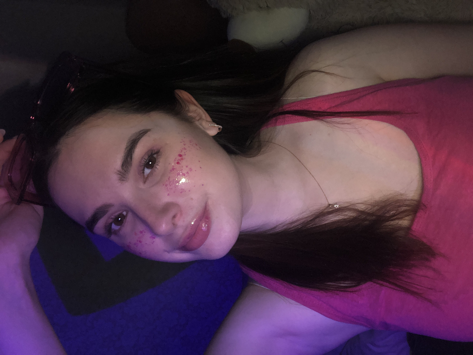 A young person, likely female, with dark hair, dark eyebrows, and brown eyes, is lying down and looking directly at the camera. She is wearing a vibrant pink tank top and has a delicate necklace with a small pendant. Her face is adorned with pink glitter, particularly around her eyes and cheekbones, adding a festive or playful touch. On her left ear, an elf ear prosthesis is visible, suggesting a costume, themed event, or personal style choice. A pair of sunglasses or eyeglasses is resting on top of her head in her dark hair.
The setting appears to be indoors, possibly a bedroom or lounge area, with dim, artificial lighting that casts a soft purple and pink hue over the scene. She is resting on a dark blue or purple surface. In the dark background, part of a light-colored plush toy or pillow is faintly discernible. The person has a slight smile, conveying a calm and engaged expression. The image captures a posed portrait with a distinct aesthetic, possibly in Noviy, Russia, as per the location context. There is no visible text in the image.