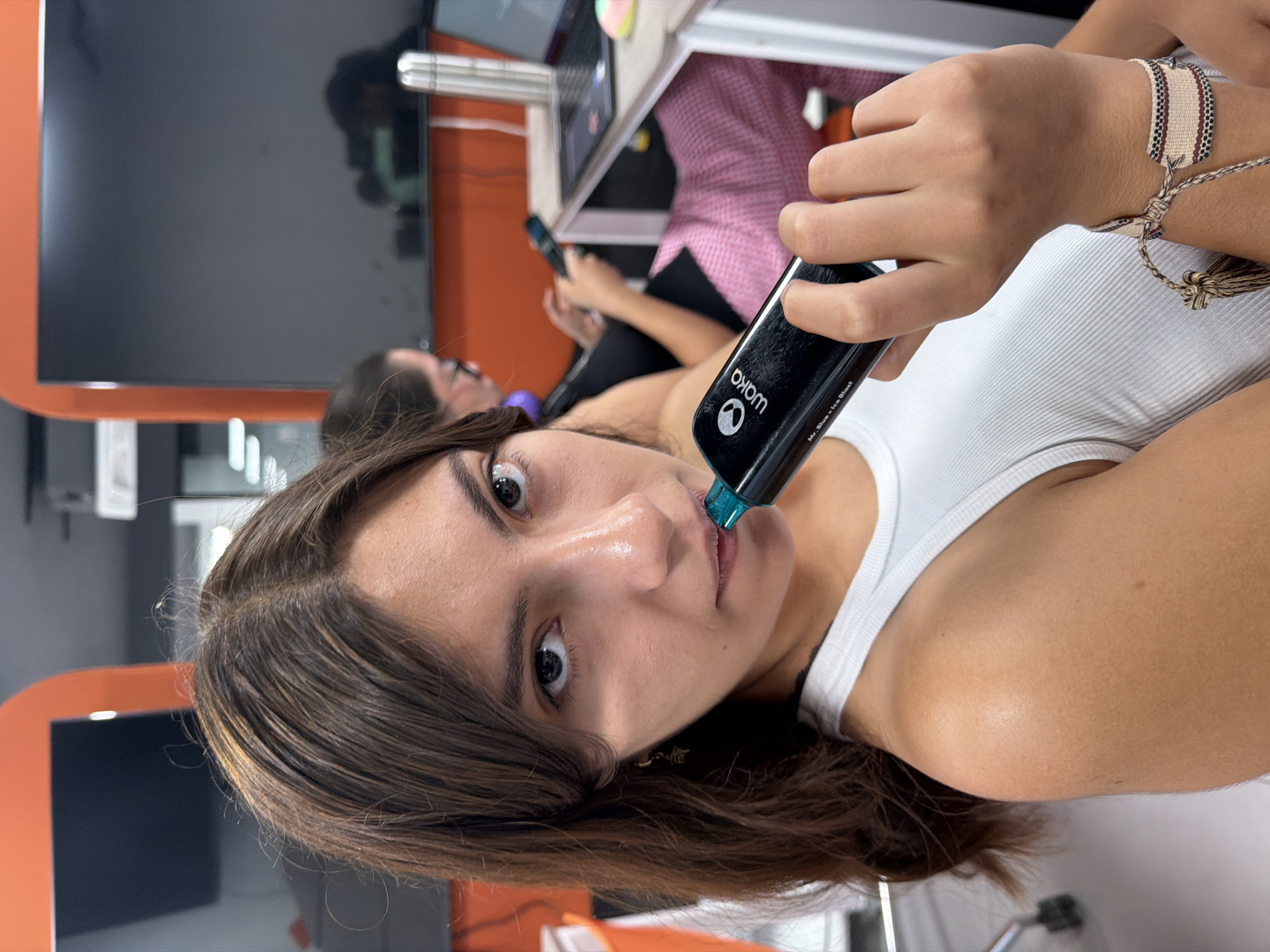 A young woman with brown hair and brown eyes is holding a black Waka vape pen with a blue tip to her lips. She is wearing a white tank top and a bracelet on her right wrist. She is indoors in a room with orange and grey walls, a monitor, and a desk with a laptop on it. Another woman is sitting in the background. The location is San Andrés Cholula, Mexico.
