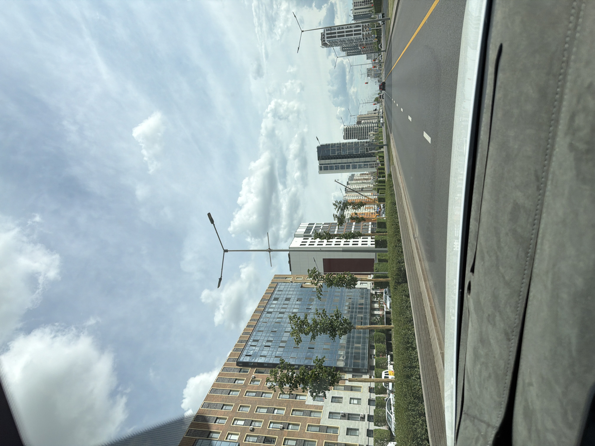 This is a vertical, eye-level, outdoor shot taken from the perspective of a vehicle moving along a multi-lane road. The road has a yellow dividing line and white lane markings. On the left side of the road are several modern, multi-story buildings with glass facades and balconies, interspersed with trees and streetlights. On the right side, a portion of the vehicle's interior, possibly a dashboard or door panel, is visible, covered in a textured gray material. The sky is cloudy with patches of blue, suggesting a partly cloudy day. The scene appears to be a contemporary urban environment, likely in Astana, Kazakhstan, given the context. The time of day appears to be daytime, likely late morning or early afternoon, given the bright, diffused light. There are no visible people in the frame. The overall impression is one of urban development and modern architecture under a dynamic sky.