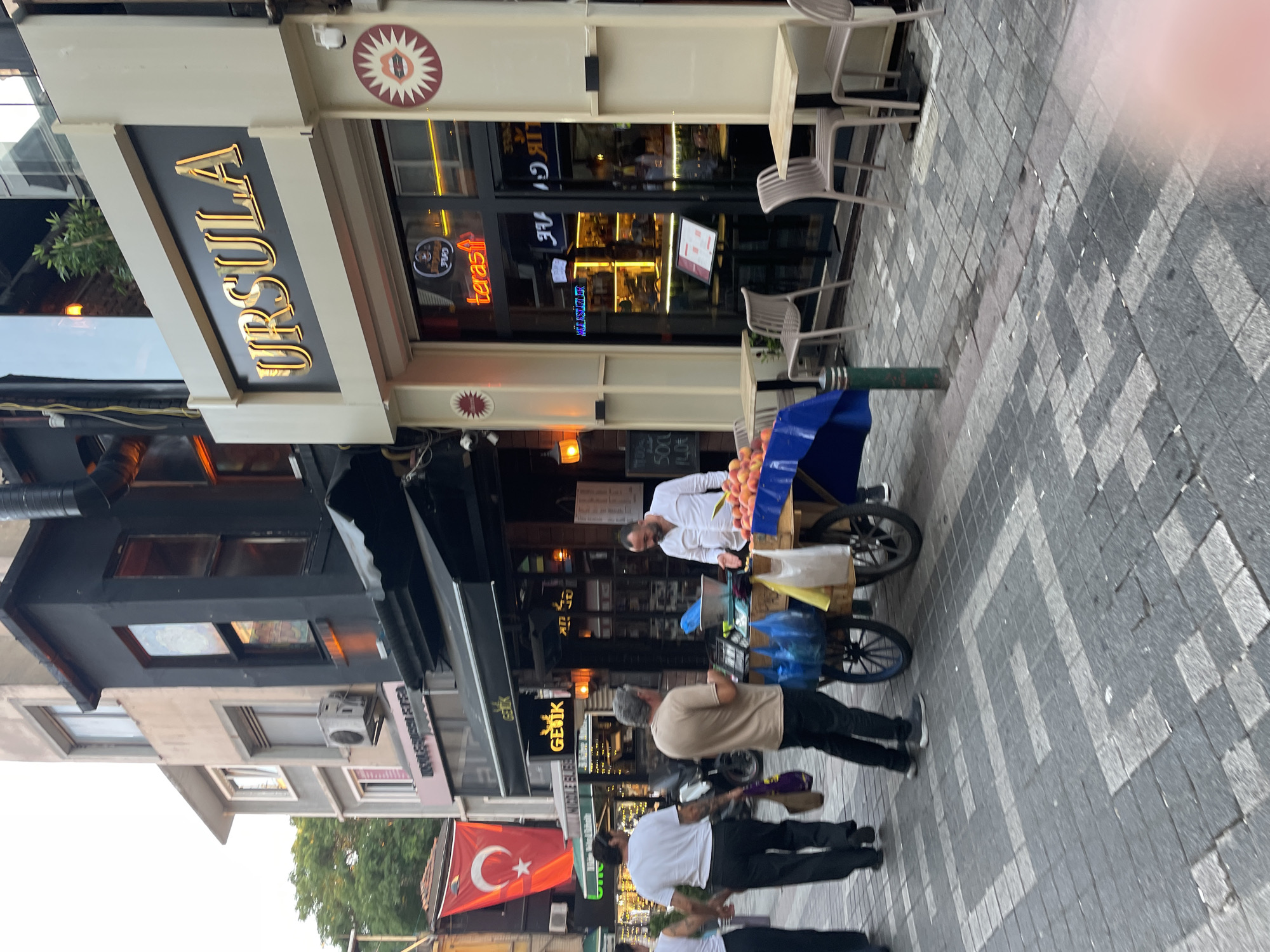 In Kadıköy, Turkey, a street scene unfolds with a fruit vendor's cart at the forefront. The cart, laden with peaches and apples, is positioned on a cobblestone street. Two men are near the cart, one man in a light brown t-shirt and black pants appears to be interacting with the vendor, who is wearing a white button-down shirt. A third man, also in black pants and a white t-shirt, walks away from the cart. The background features a storefront with the prominent signage "URSULA" in gold lettering. Additional storefronts and buildings line the street, including one with a Turkish flag hanging outside. The environment suggests an urban setting with pedestrian traffic and commercial establishments.