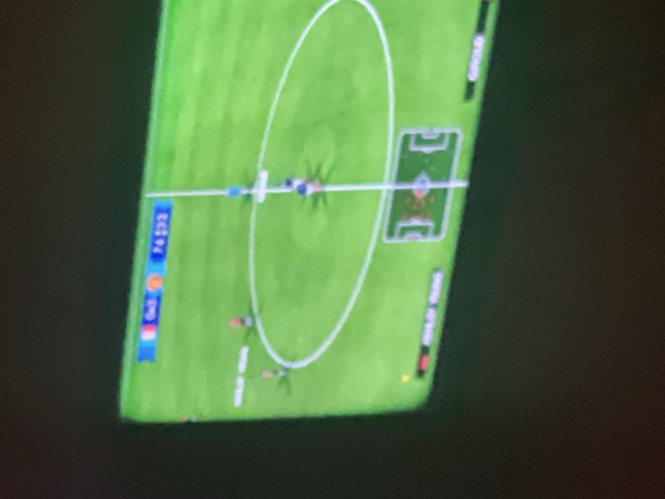 A soccer game is shown from a top-down perspective. A bright green field is marked with white lines, including a center circle and goal areas. Several miniature figures, presumably representing soccer players, are scattered across the field. A white ball is positioned in the center circle. Scores and team colors are visible along the left and right edges of the screen.
