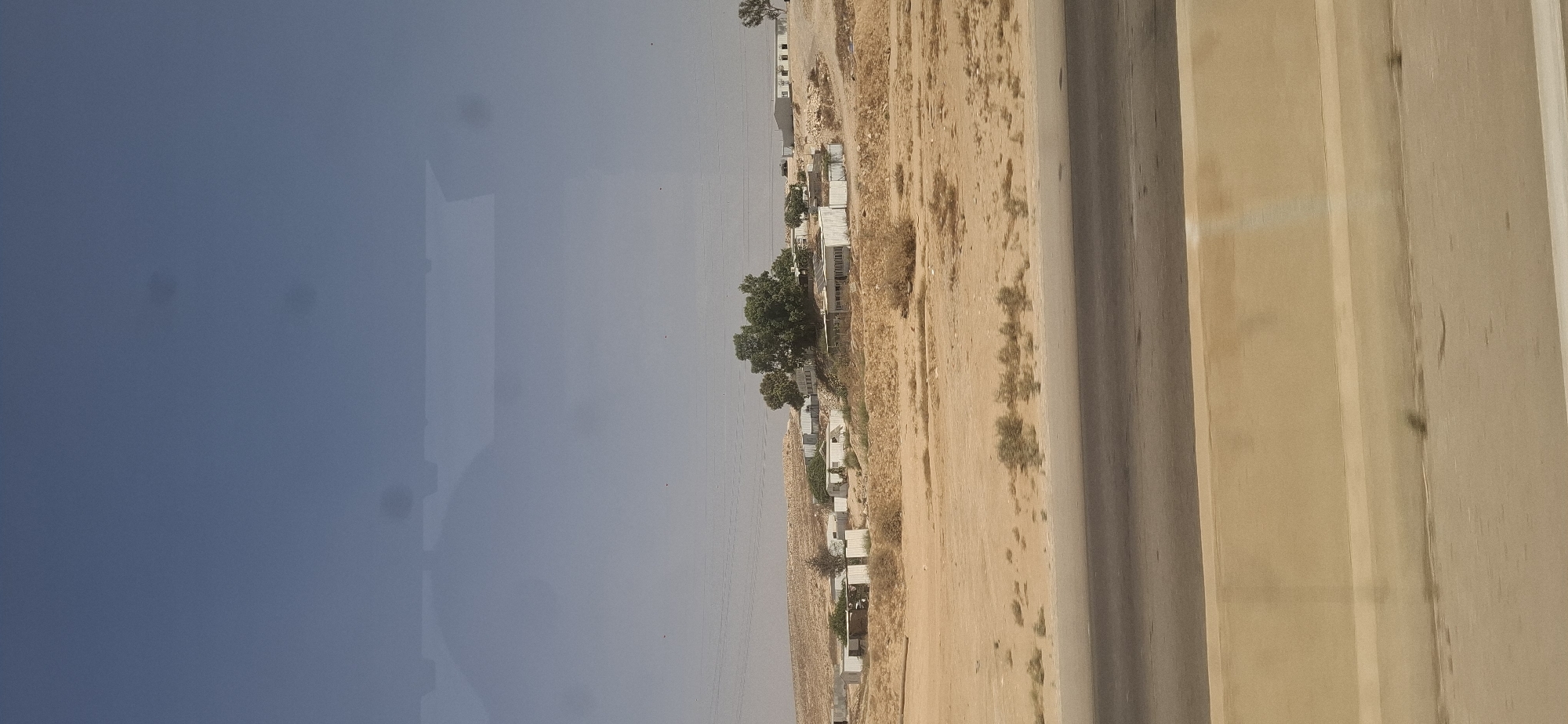 This is a photograph of a desert landscape in the Merhavim Regional Council, Israel. In the background, there are several small, white buildings clustered on a hillside, some with solar panels visible on their roofs. A small grove of green trees stands behind some of the buildings. The land is dry and sandy, with scattered sparse vegetation. In the foreground, a grey asphalt road runs horizontally across the bottom of the frame, separated from the desert by a concrete barrier. The sky is a hazy light blue. There are no people visible in the image. The overall impression is one of arid rural settlement.