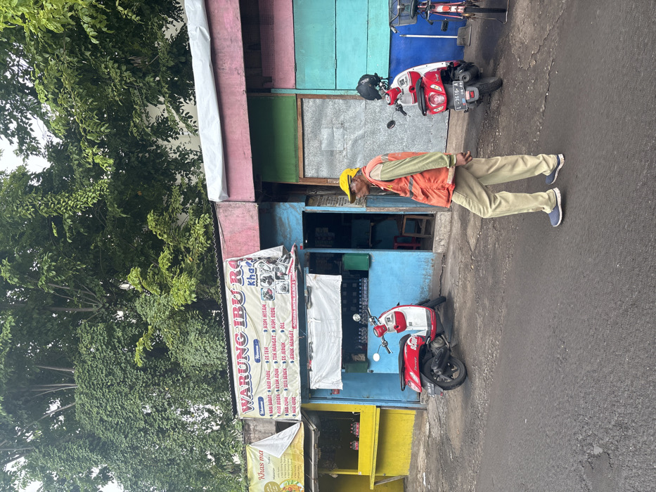 A man in a yellow hard hat and orange vest stands near a colorful roadside shop called "Warung Ibu Ra," offering items like coffee, tea, and juice. The shop is painted with various bright colors and banners advertising its offerings. Two parked red and white motorbikes sit near the entrance, with one possibly obscuring part of the shop. Trees provide shade along the side of the street.