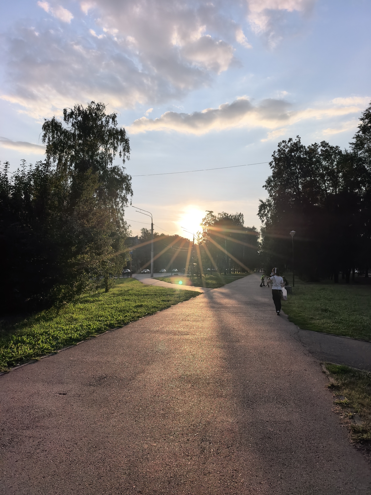 An outdoor, low-angle, eye-level, full shot shows a paved path in a park at sunset in Sterlitamak, Russia. The sun is low on the horizon and beams outward, creating long shadows on the path. The path is lined with green grass and trees on both sides. A lone individual, seen from behind and walking away from the camera, is on the path. They appear to be carrying a white bag in their right hand and are dressed in dark clothing. Cars are visible in the background, and a lamppost stands to the right of the path. The sky is partly cloudy, with some of the clouds illuminated by the setting sun.