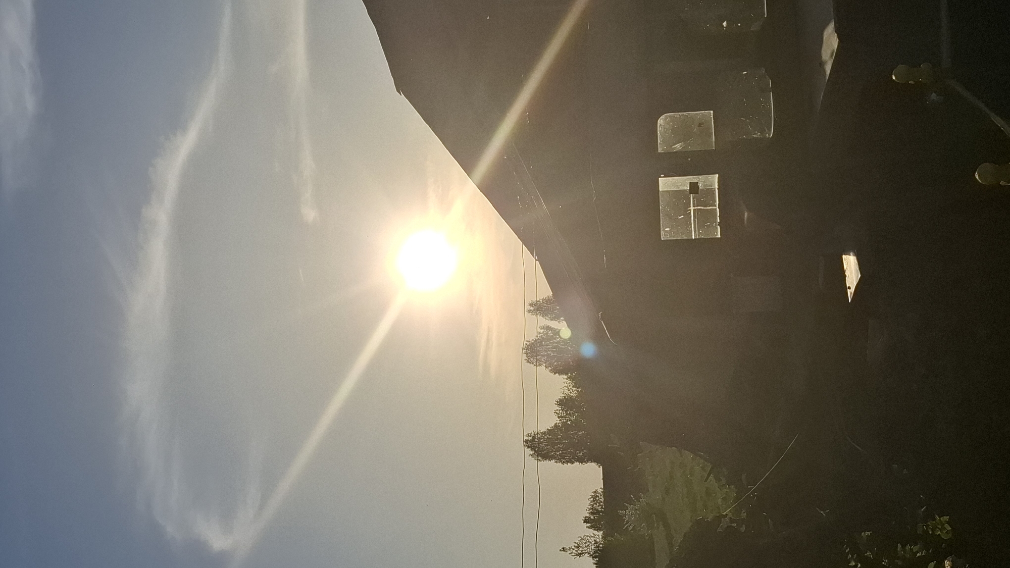 The image shows a scene with a bright sun in the sky. There are thin clouds visible. To the right of the sun, there appears to be a building or structure, possibly with windows. Trees and greenery are visible along the bottom edge of the image. There is no evidence to suggest it is in the city of Шешурга, Russia. The image primarily depicts natural elements (sky, sun, clouds, trees) and a structure, possibly a building.