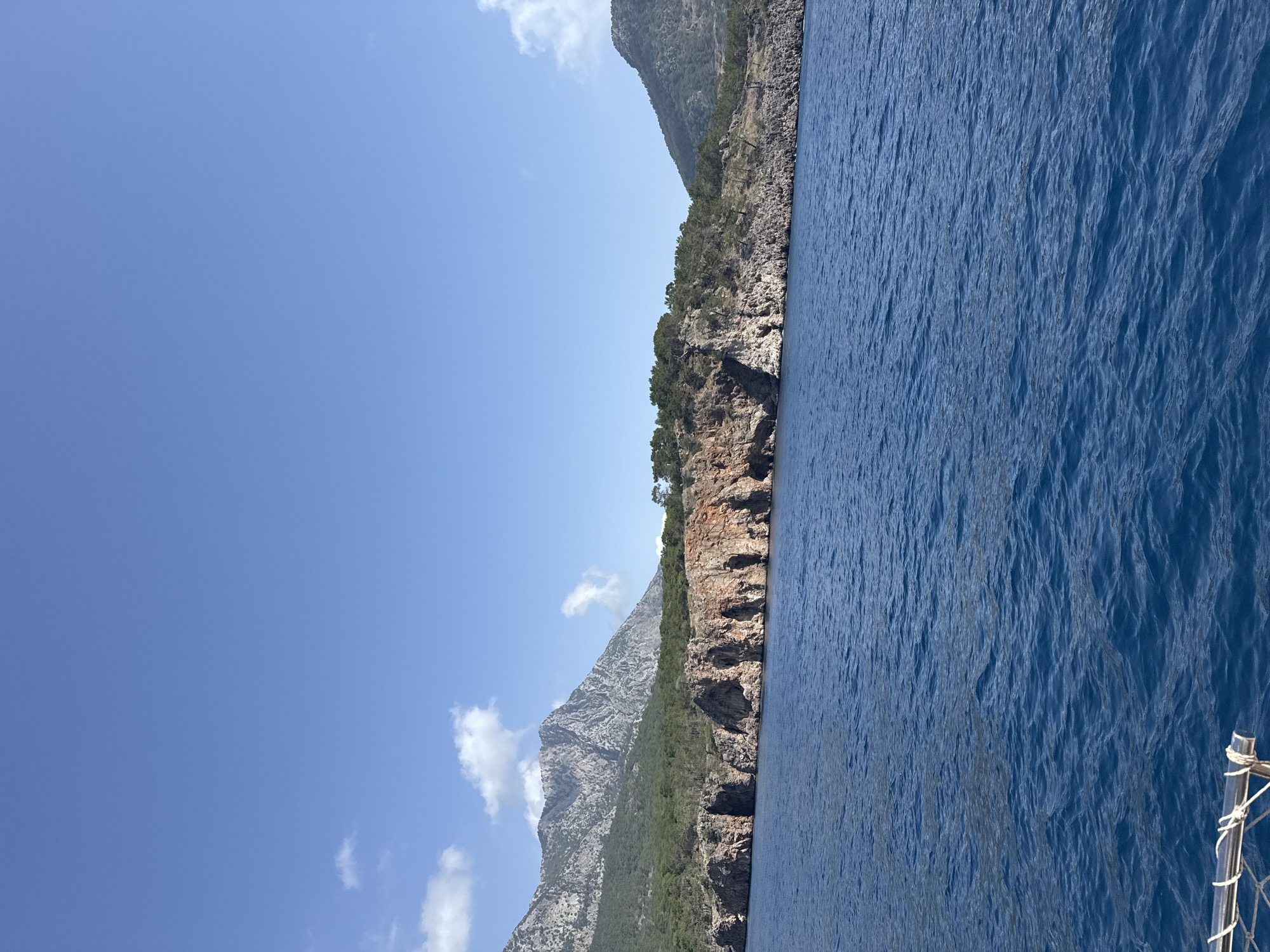 The image shows a coastline with rocky cliffs covered in vegetation, next to deep blue water. The sky above is clear and blue with a few white clouds. Parts of a boat are visible on the bottom right.
There is no evidence that this photo was taken in Unknown, Türkiye.