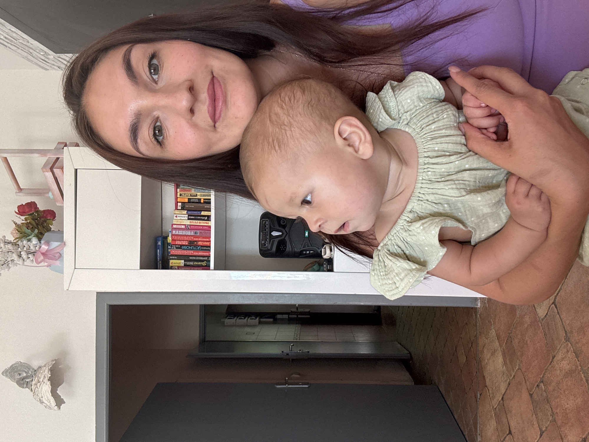 A woman with long, dark brown hair and fair skin is holding a baby. The woman is wearing a purple top. The baby is wearing a light green, ruffled romper. The baby is looking to the left of the frame. Behind the woman, a white bookshelf is visible, filled with books and a black speaker. To the left of the bookshelf is a doorway leading to another room with a tiled floor. The overall setting appears to be an indoor residential environment.