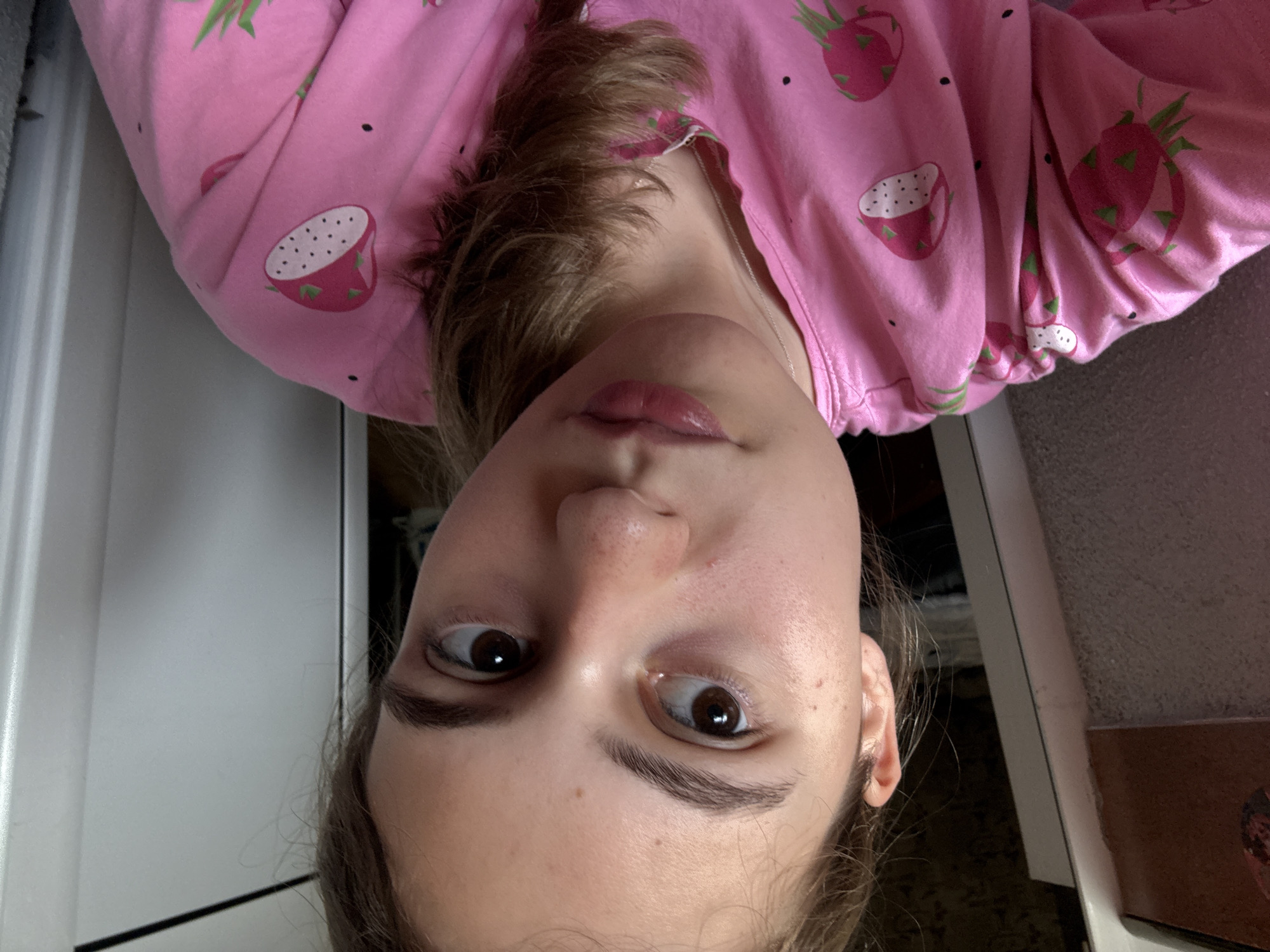 A young woman with light brown hair and brown eyes is shown in a close-up, selfie-style shot. She is wearing a pink pajama top adorned with a pattern of strawberries and dragon fruit. Her hair is pulled back into a ponytail and cascades over her right shoulder. The background is softly blurred, showing elements of a room, including a white doorframe and a dark, textured surface on the left. The lighting appears to be natural, possibly from a window. The image was taken indoors. There is no information available about the specific location in the city of Panagyurishte, Bulgaria.