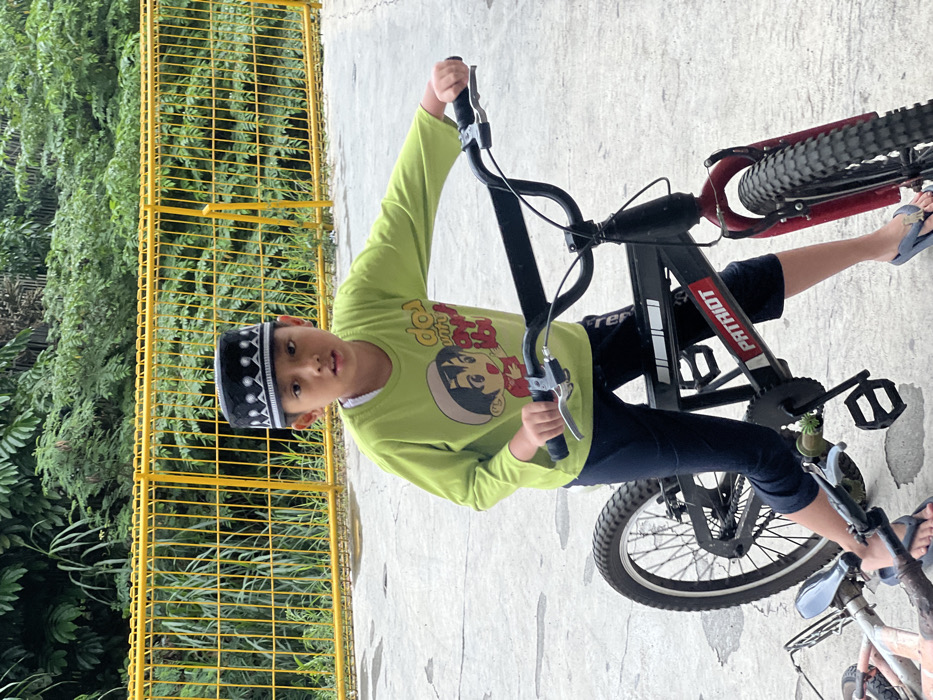 A young male, appearing approximately 7-9 years old, stands centrally positioned, straddling a black BMX-style bicycle on a light gray concrete surface. He wears a black peci (songkok) adorned with white geometric embroidery, a light green long-sleeved shirt featuring a multi-color cartoon character graphic and text, dark blue cropped pants, and blue flip-flops. His hands grip the black handlebars of the bicycle, which has a red front fork and knobby tires. The bicycle frame prominently displays the word "PATRIOT" in white lettering. The subject's gaze is directed forward, towards the viewer. In the background, a bright yellow painted metal fence runs horizontally, behind which dense green foliage from various plants is visible. A portion of a smaller, light-colored object, possibly another bicycle part, is visible on the ground to the lower left. The scene depicts an outdoor setting, consistent with a residential or recreational area within a city such as Kecamatan Pati, Indonesia.