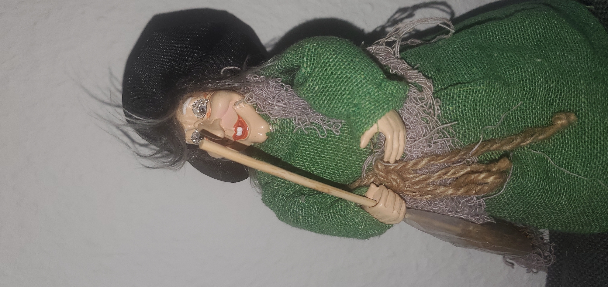 A close-up, eye-level, medium shot captures a doll representing a witch. The doll has a light-colored face with a large, pointed nose, rosy cheeks, a wide red smile, and exaggerated eyelashes decorated with glitter. Its grey hair is wispy and appears windblown. It wears a black, wide-brimmed hat and a coarse, green woven dress. A light purple, frayed scarf is draped around its neck. The doll holds a light-brown wooden stick in its right hand, with a thick, braided brown rope wrapped around its middle and the stick. Its left hand is slightly open, positioned near the rope. The background is a plain, textured light gray wall, with a shadow cast by the doll on the right side. The doll is positioned indoors, and the scene is static. The specific location in the city of Sasbach, Germany is not discernible from the image.
