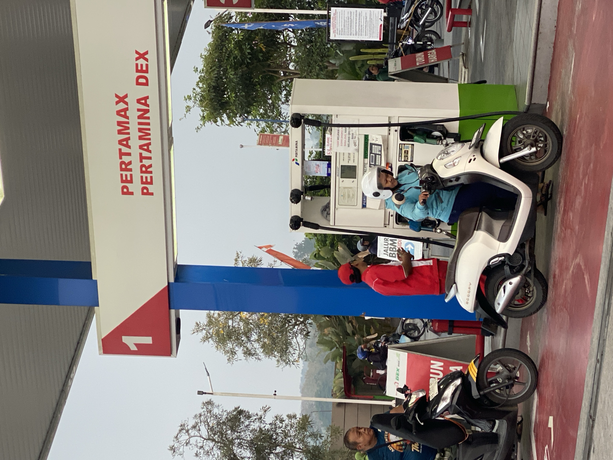 The image shows a gas station in Kasihan, Indonesia. The gas station has multiple pumps, and several people are present, including a woman on a white scooter wearing a helmet and a man in a red uniform holding a sign that says "JALUR BBM MASUK". There are also other scooters and motorcycles parked at the station. The signage indicates that the station sells Pertamax and Pertamina Dex fuel. The environment appears to be an outdoor scene with trees and greenery in the background.