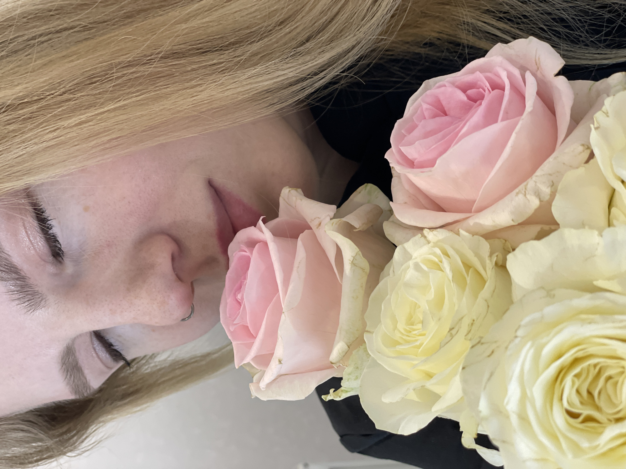 A close-up, slightly tilted image shows a fair-skinned person with blonde hair next to a bouquet of roses. The person has visible freckles, a nose piercing, and their eyes are closed or looking down. The bouquet contains roses in varying shades of pink and cream. A dark garment or background is visible behind the flowers. The image does not contain enough information to identify a location in Izhevsk, Russia, or to determine any actions or interactions.