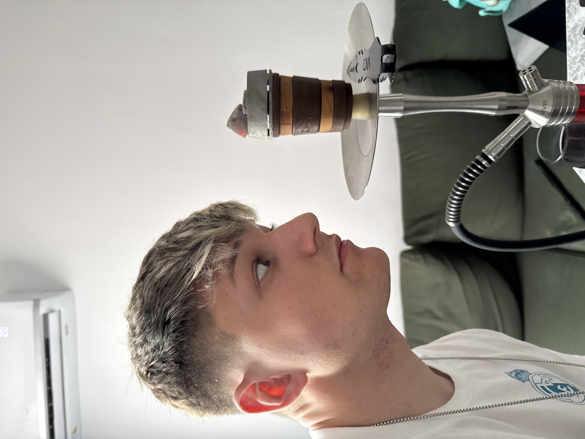 A young, light-skinned male is looking upwards at a hookah, which is set up on a table in the background. The hookah has a lit coal. The man is wearing a white t-shirt with a silver chain. In the background is a green couch and a white air conditioning unit. The location is not discernable as being in Plovdiv, Bulgaria.