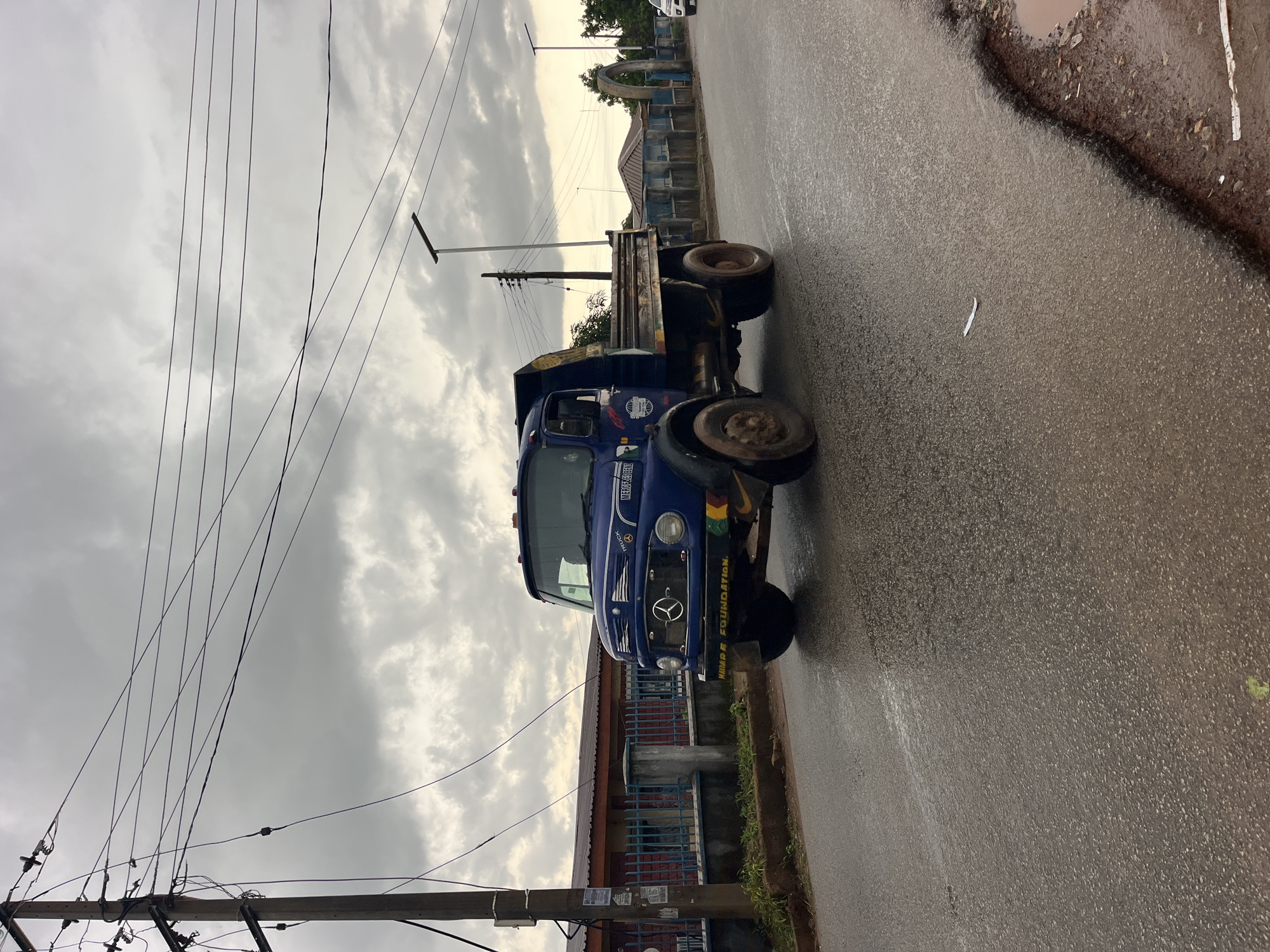 A blue truck is parked on a wet asphalt road, which appears to be slightly damaged with a puddle of water on the right side of the frame. The truck is facing towards the right of the image. It has a Mercedes-Benz logo on the front grill. The side of the truck has some writing on it, which appears to say "HMP FOUNDATION". The sky above is overcast and cloudy. There are power lines visible in the upper left of the image, running diagonally. In the background, there are some buildings with what looks like blue fencing or railings. The overall lighting suggests it could be daytime, possibly late afternoon or early morning given the cloud cover. The wet road indicates recent rain or ongoing damp conditions. There are no people visible in the image. The scene appears to be in an urban or semi-urban environment.