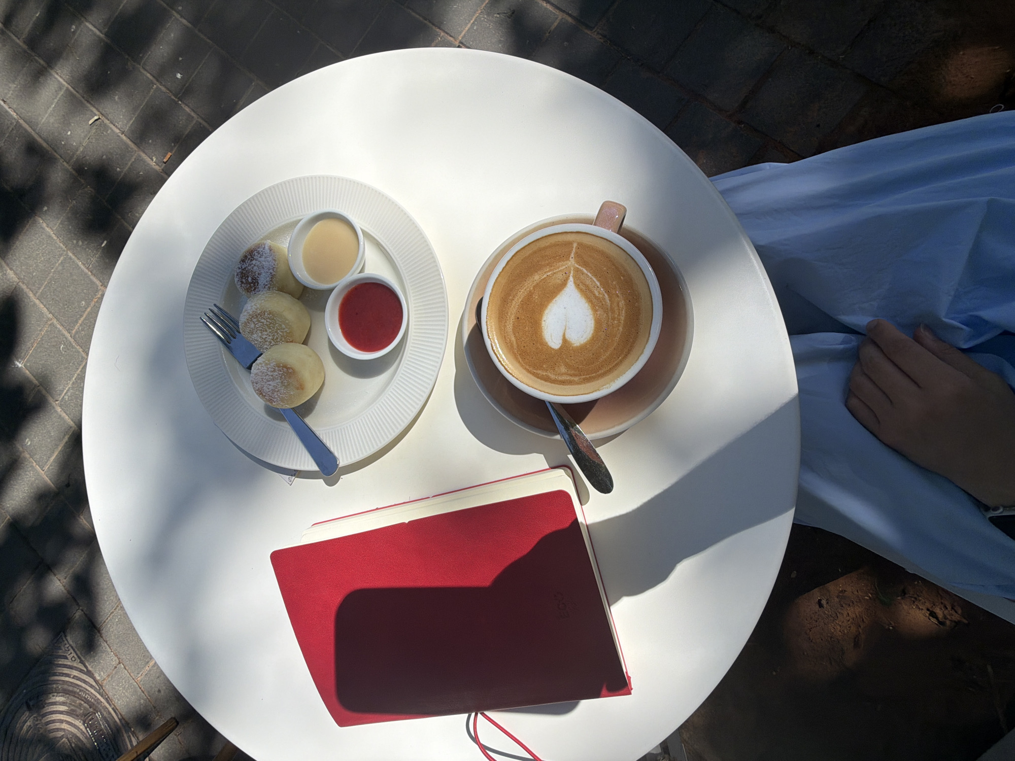 The scene depicts an outdoor cafe setting in Bat Yam, Israel, on a sunny day with strong directional light creating prominent shadows. A person is seated at a white, circular table, with only their lower body and right hand visible. They are wearing a light blue garment, possibly a dress or skirt, with their hand resting on their lap.
On the table, positioned from top to bottom:
1. A white ceramic plate with a fluted rim holds three small, round, baked items, resembling syrniki or small pancakes, dusted with powdered sugar. A small, light blue-handled fork is placed on the plate.
2. Adjacent to the plate, two small white ramekins contain sauces: one with a vibrant red, thick liquid (likely a berry jam), and the other with a pale yellow, creamy liquid (possibly condensed milk or custard).
3. A light pink/beige ceramic coffee cup with a matching saucer holds a beverage featuring distinct heart-shaped latte art. A silver spoon rests beside the cup.
4. To the left, a red hardcover notebook or journal is visible, partially open, with a red elastic band. A rectangular shadow, consistent with a mobile phone, is cast across its cover.
The ground surrounding the table is paved with dark grey bricks, and overhead foliage casts dappled shadows across the scene.