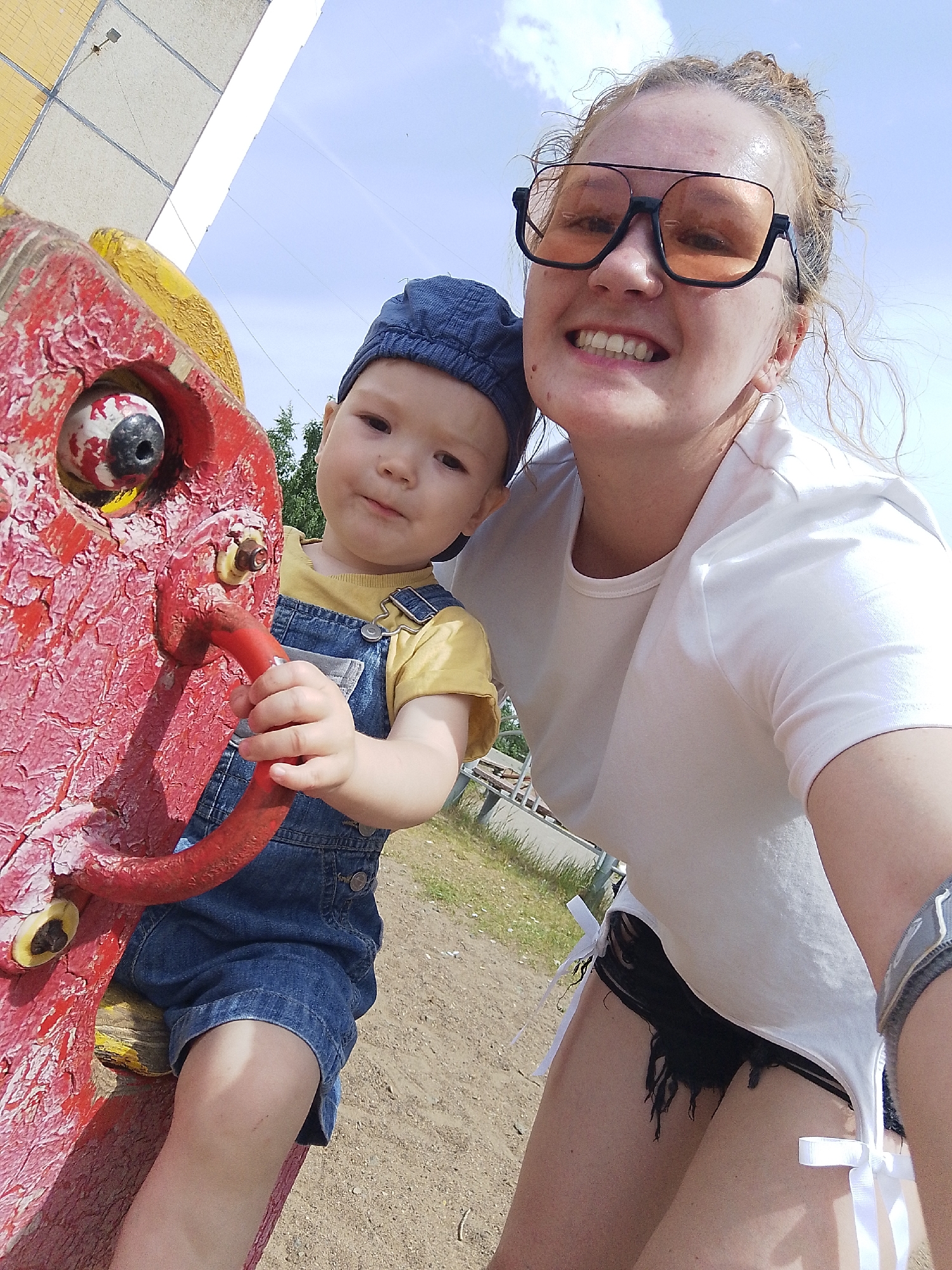 A photograph captures a woman and a young child next to a weathered, red-painted playground structure with a cartoonish horse head. The image, presumably taken in a park or playground in Chaikovsky, Russia, presents a sunny outdoor setting.
The woman, positioned on the right, wears large, black-rimmed sunglasses with amber lenses and a white T-shirt over black shorts. She smiles broadly at the camera, revealing her teeth. Her light-colored hair is pulled back.
The child, to the left, is seated on the red playground structure, which features a prominent, discolored eye on the horse head. The child is dressed in denim overalls over a yellow shirt and a blue cap. He holds onto a red handlebar.
The background shows parts of the building and some green trees. The ground around the playground is sandy. The weathered condition of the playground structure suggests it has been exposed to the elements for an extended period.