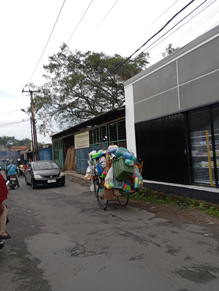 A street scene in Kecamatan Kutawaringin, Indonesia (latitude: -6.974, longitude: 107.547). A person in red clothing is walking on the street, facing towards the left side of the image. A person is riding a motorcycle on the street, facing towards the left. A grey car is stopped in the middle of the street, facing towards the left. A motorcycle is parked on the left side of the street. A cart loaded with various objects including a plastic bucket, a broom, and other household items, is being pulled by a man on the right side of the street. The man is obscured from view. There are power lines running across the top of the image. A building with a black metal door is on the right side of the image. A tree is on the left side of the image, with the top of the tree branches out of frame. The ground is paved asphalt, and the street has cracks and potholes. The sky is overcast and grey.